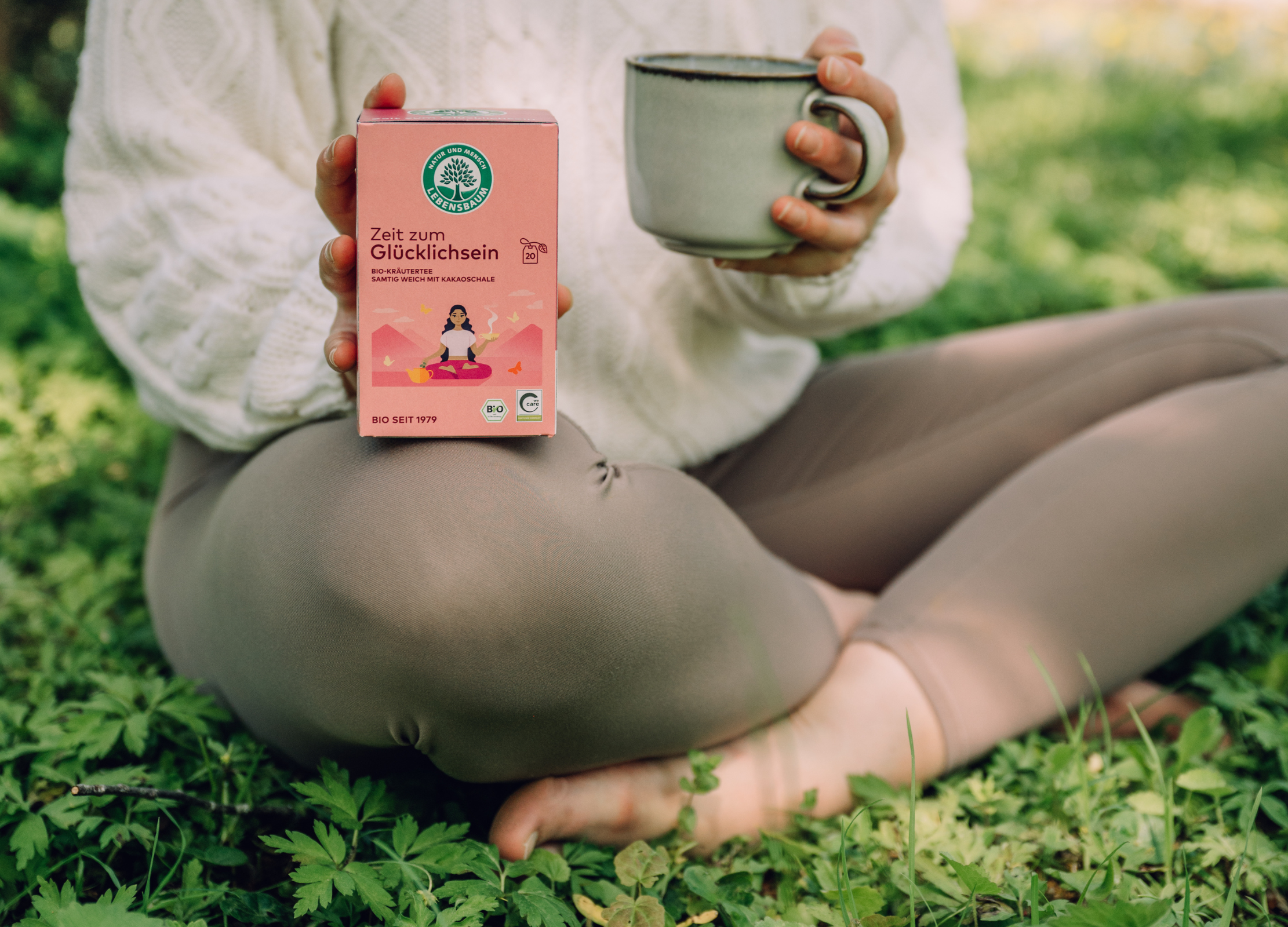 Person sitzt barfuß im Gras, hält eine Zeit zum Glücklichsein Teepackung mit Yogamotiv und eine Tasse in den Händen