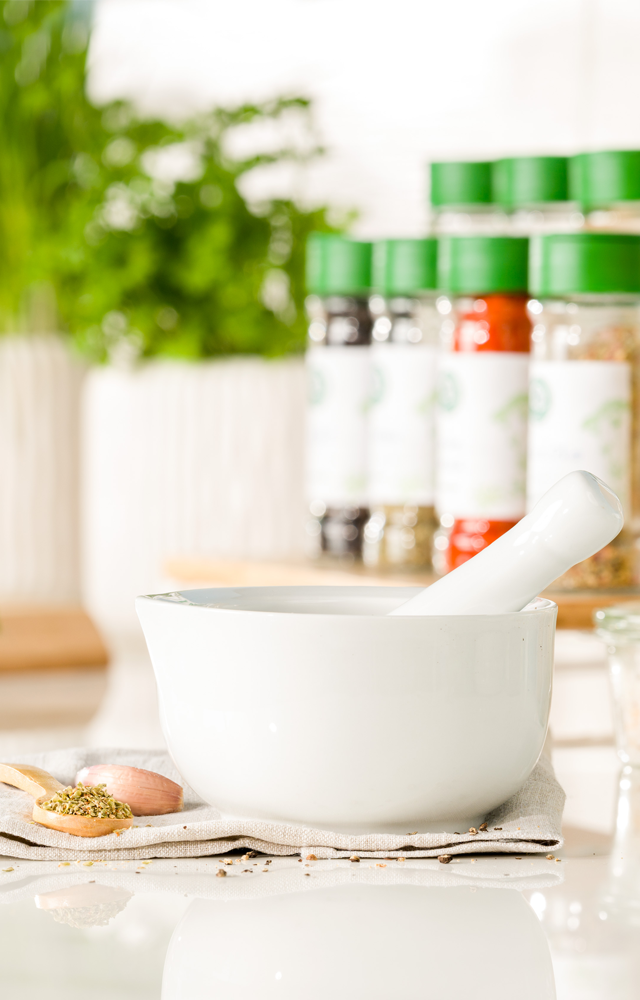 Weißer Mörser mit Stößel steht auf einem Tuch, daneben ein kleiner Löffel mit Gewürzen – im Hintergrund verschiedene Gewürzstreuer mit grünem Deckel