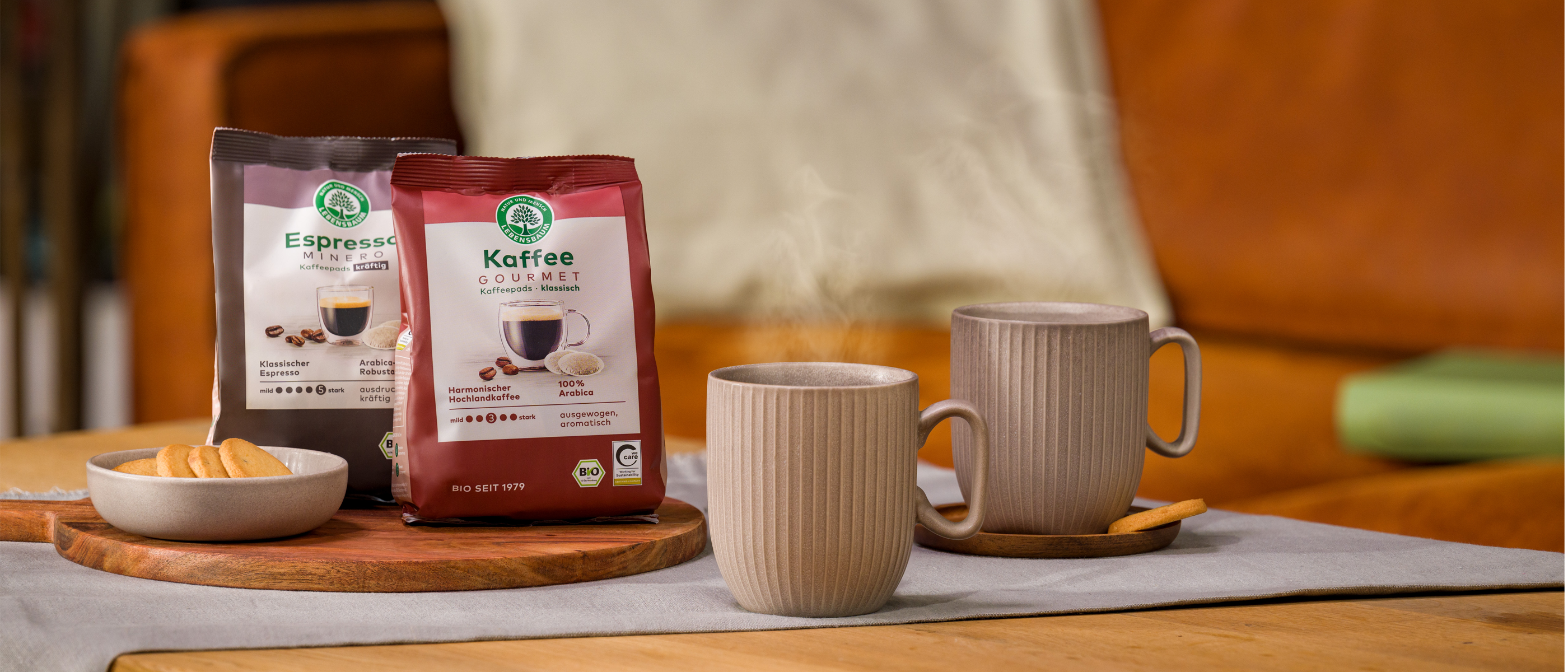 Zwei dampfende Tassen Kaffee stehen auf einem gedeckten Tisch neben einer Keks­schale und zwei Packungen Lebensbaum Kaffee und Espresso