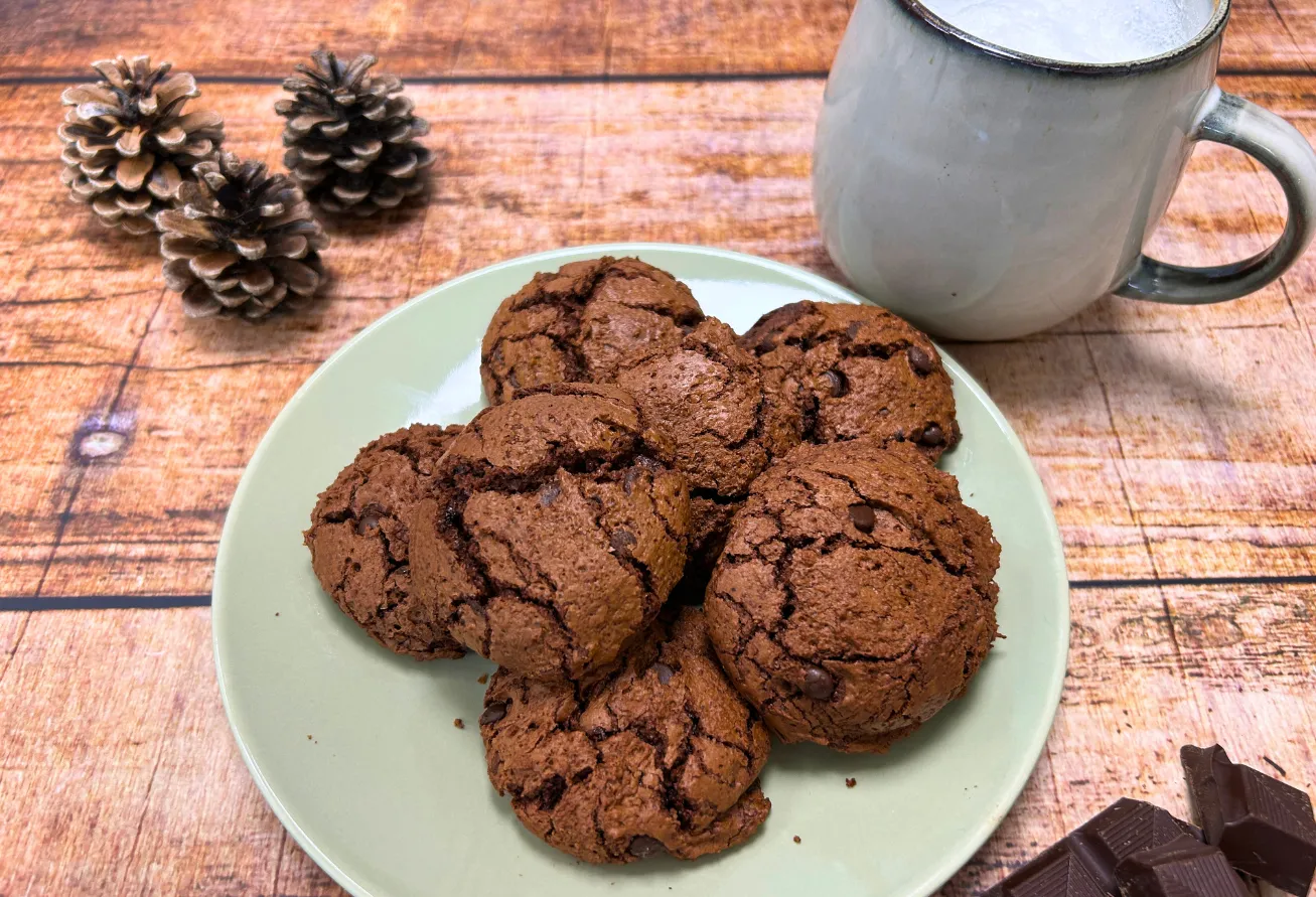 Espresso-Brownie-Cookies auf Porzellanteller mit Emaillebecher, Tannenzapfen und Schokolade auf Holztisch