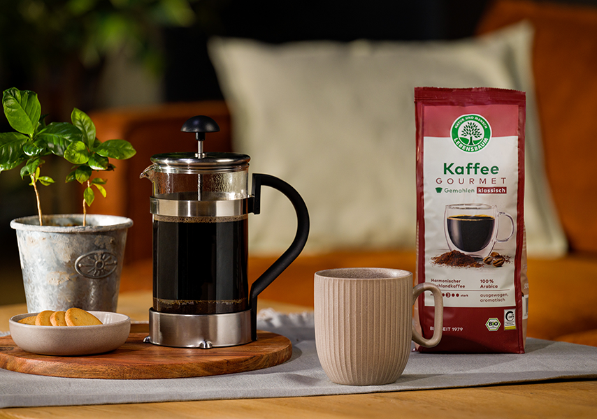 Eine Packung Lebensbaum Gourmet Kaffee steht auf einem Tisch neben einer French Press Maschine und einer Tasse