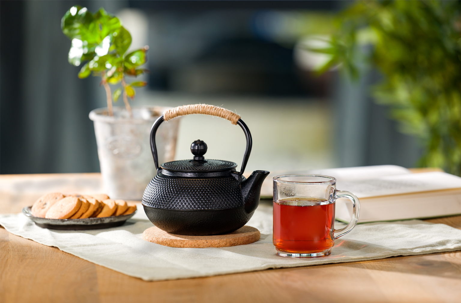 Eine schwarze Teekanne mit geflochtenem Griff steht auf einem runden Untersetzer, daneben ein Glas mit kräftigem Tee und ein Teller mit Keksen auf einem hellen Tischläufer
