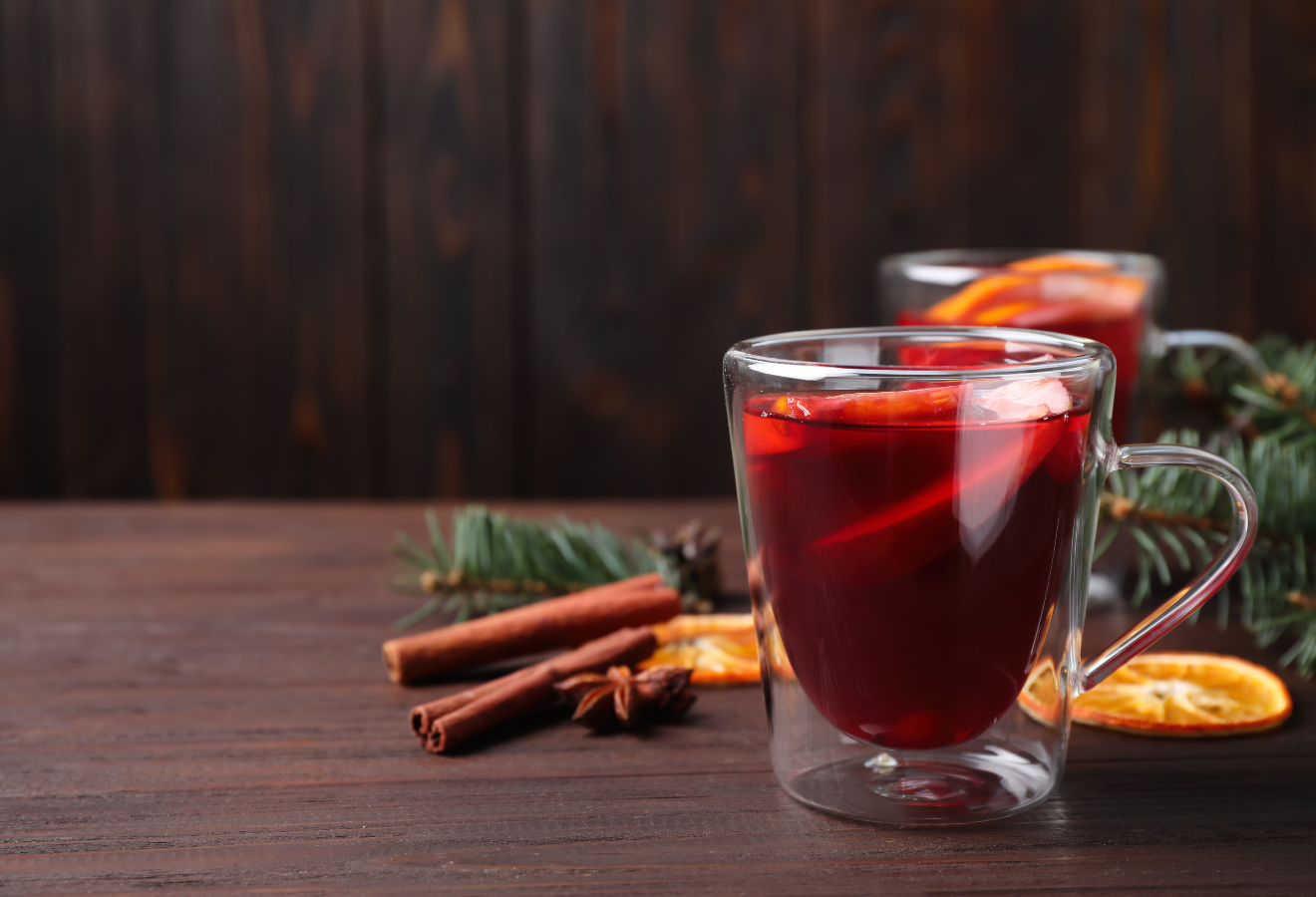 Roter Glühwein in einer Glastasse, dahinter Zimtstangen, getrocknete Orangenscheiben und Tannenäste