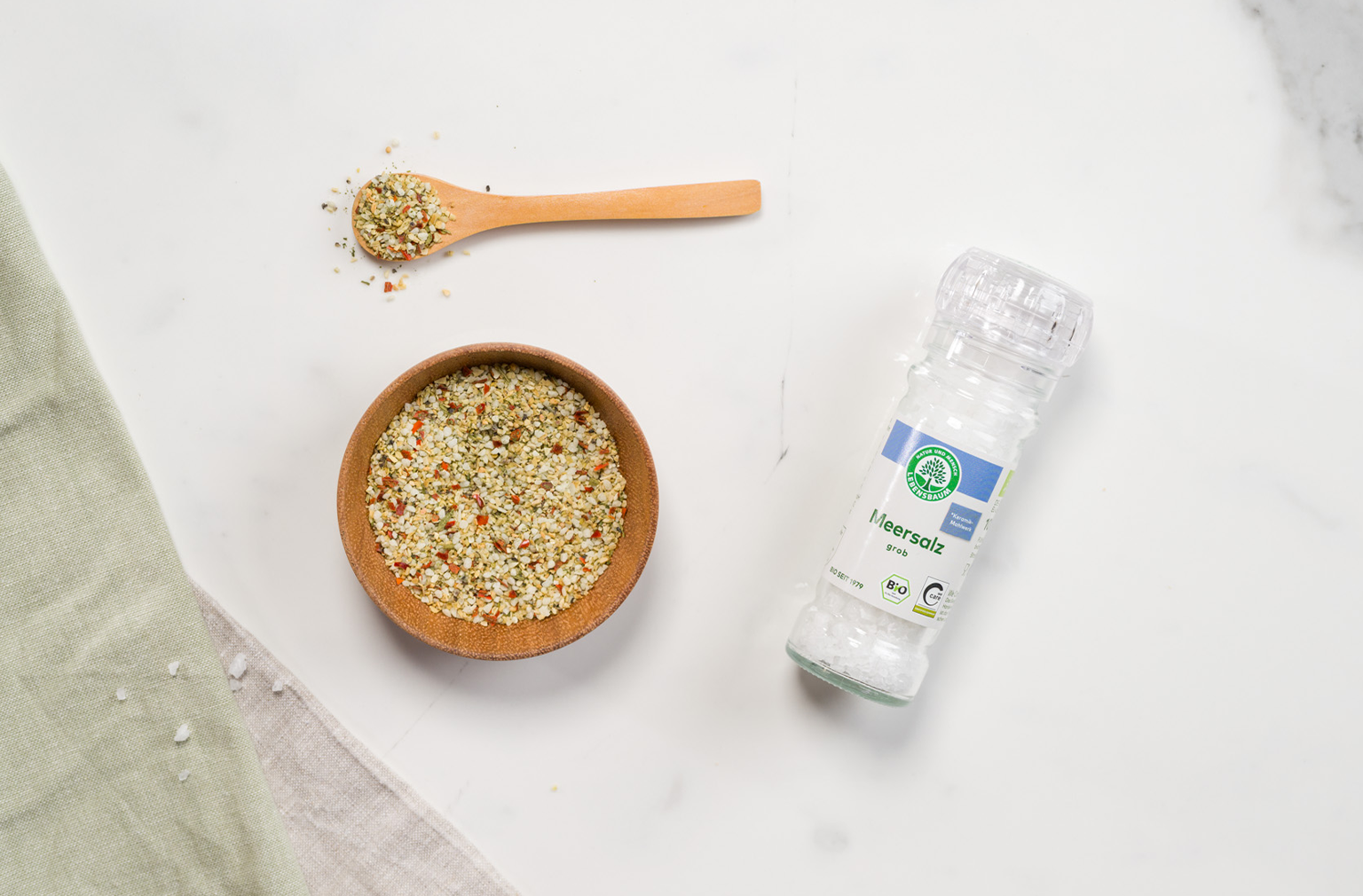 Eine Lebensbaum Salzmischung in der Mühle liegt neben einem Holzlöffel und einer Schale mit Gewürzsalz auf weißer Marmorplatte