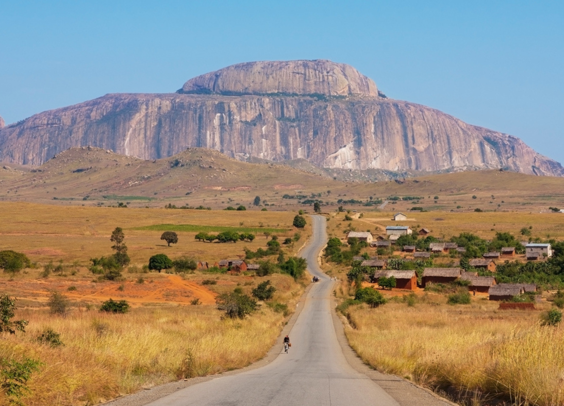 Straße führt durch Savanne auf ein Dorf zu, mit großem Tafelberg in Madagaskar im Hintergrund