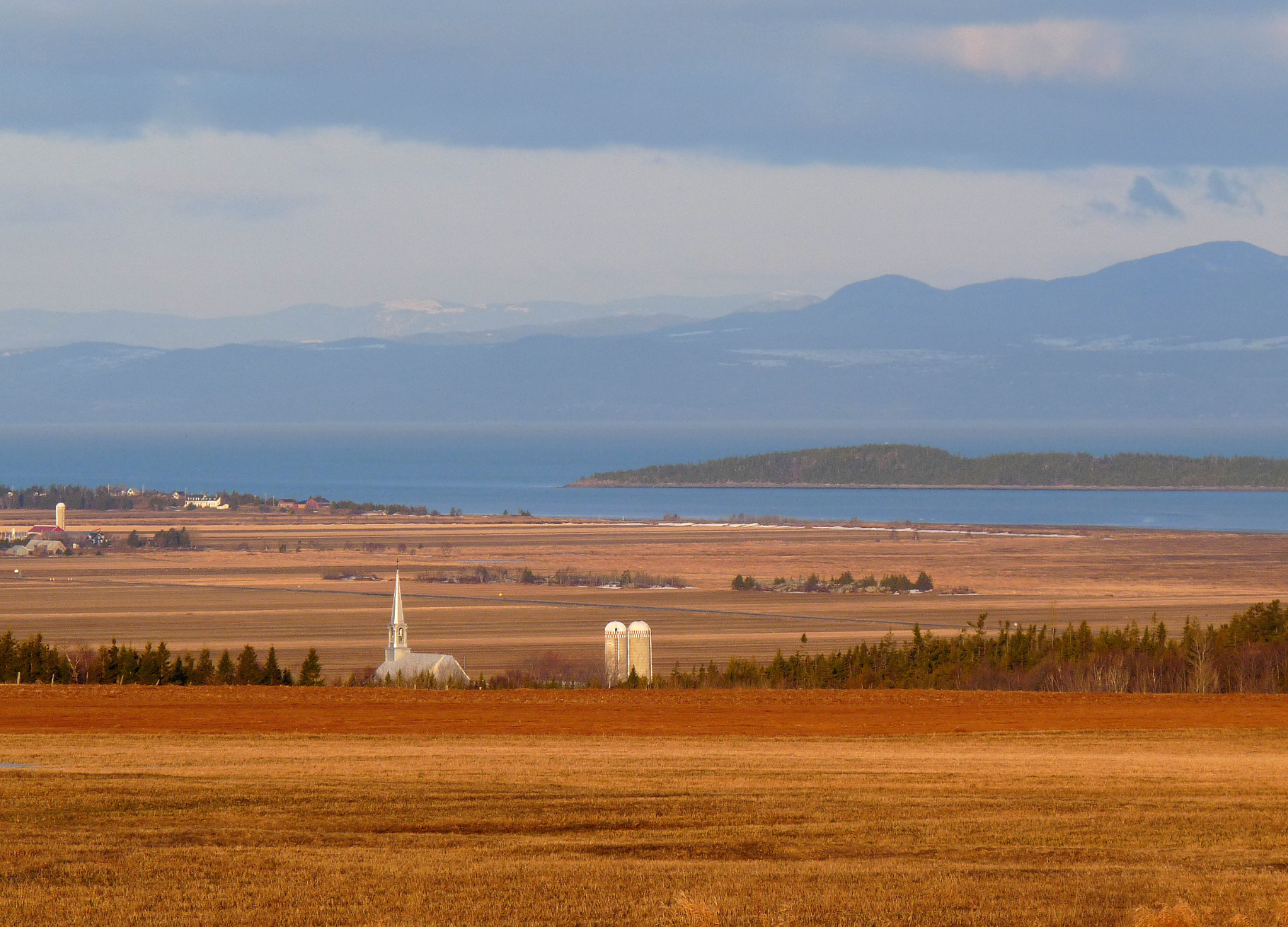 Weite Landschaft mit Feldern, einem weißen Kirchturm und Silos, dahinter ein Fluss und Berge am Horizont in Kanada
