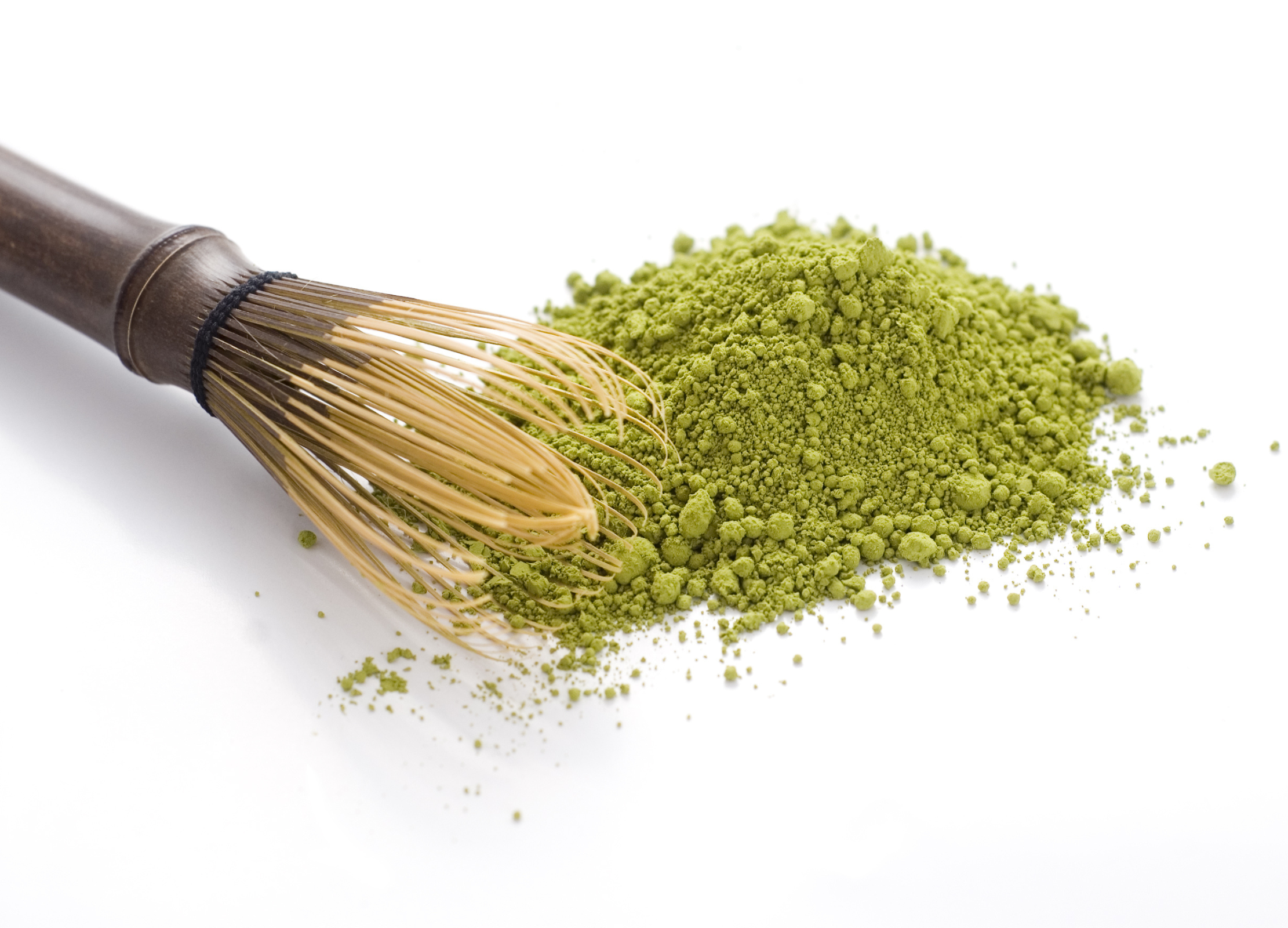 Grünes Matcha-Pulver liegt auf weißem Untergrund, daneben ein traditioneller Bambusbesen zum Aufschlagen des Tees