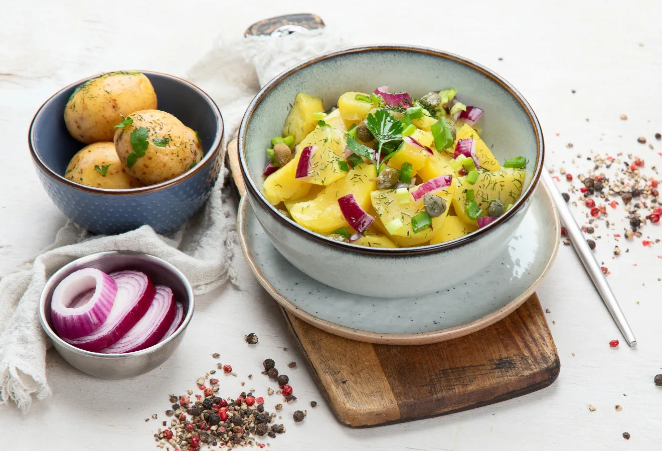 Herbstlicher Kartoffelsalat in einer Schüssel, daneben 2 Schälchen mit Zwiebelscheiben und Kartoffeln darin