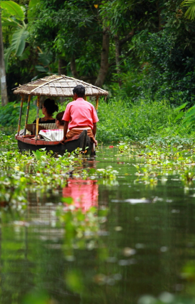 Menschen in einem kleinen Boot mit Strohdach fahren durch einen grünen Wasserkanal in Kerala