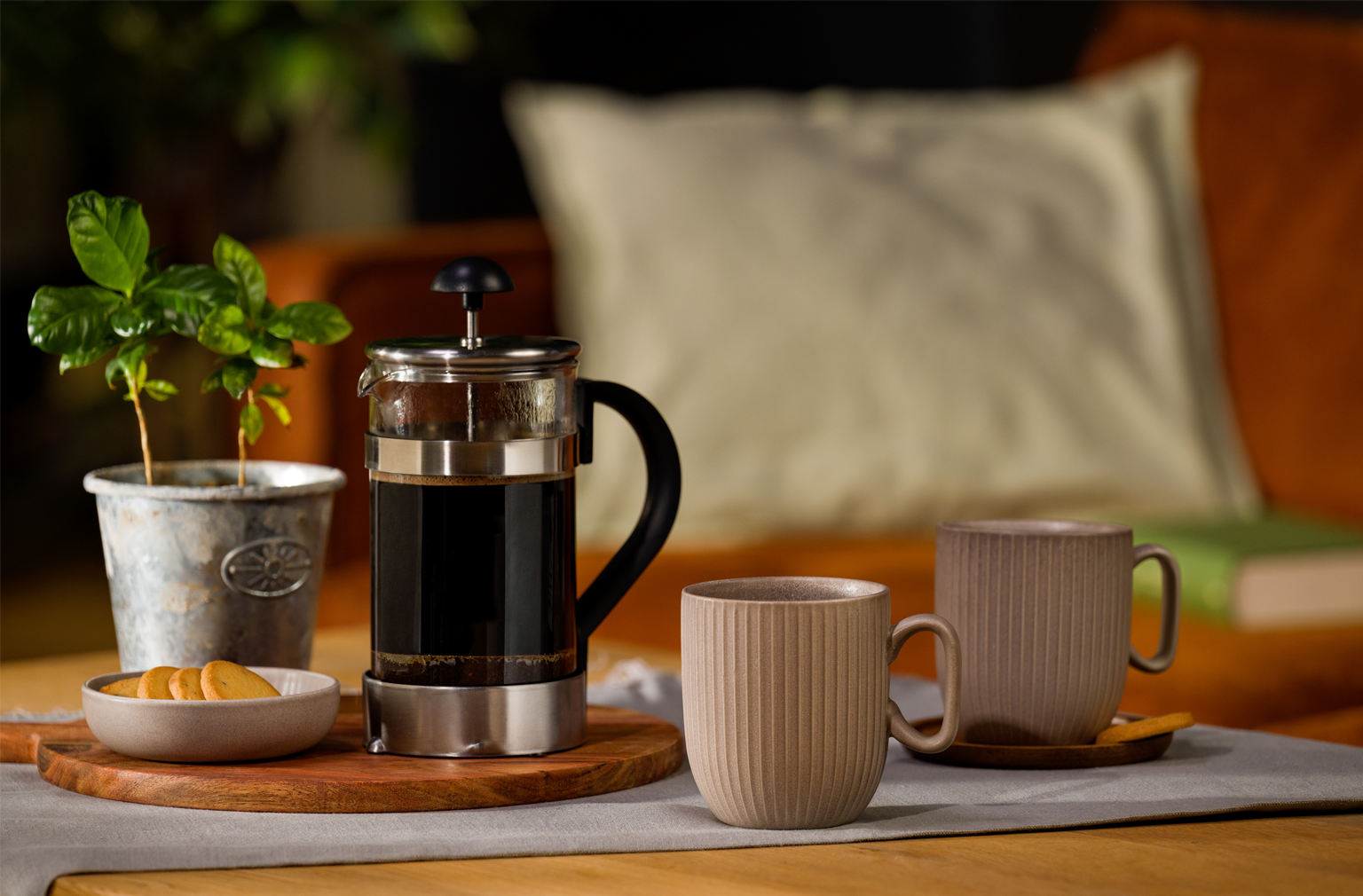 Zwei beige Tassen stehen neben einer gefüllten French Press auf einem gedeckten Tisch, daneben ein kleiner Keksteller und eine Topfpflanze