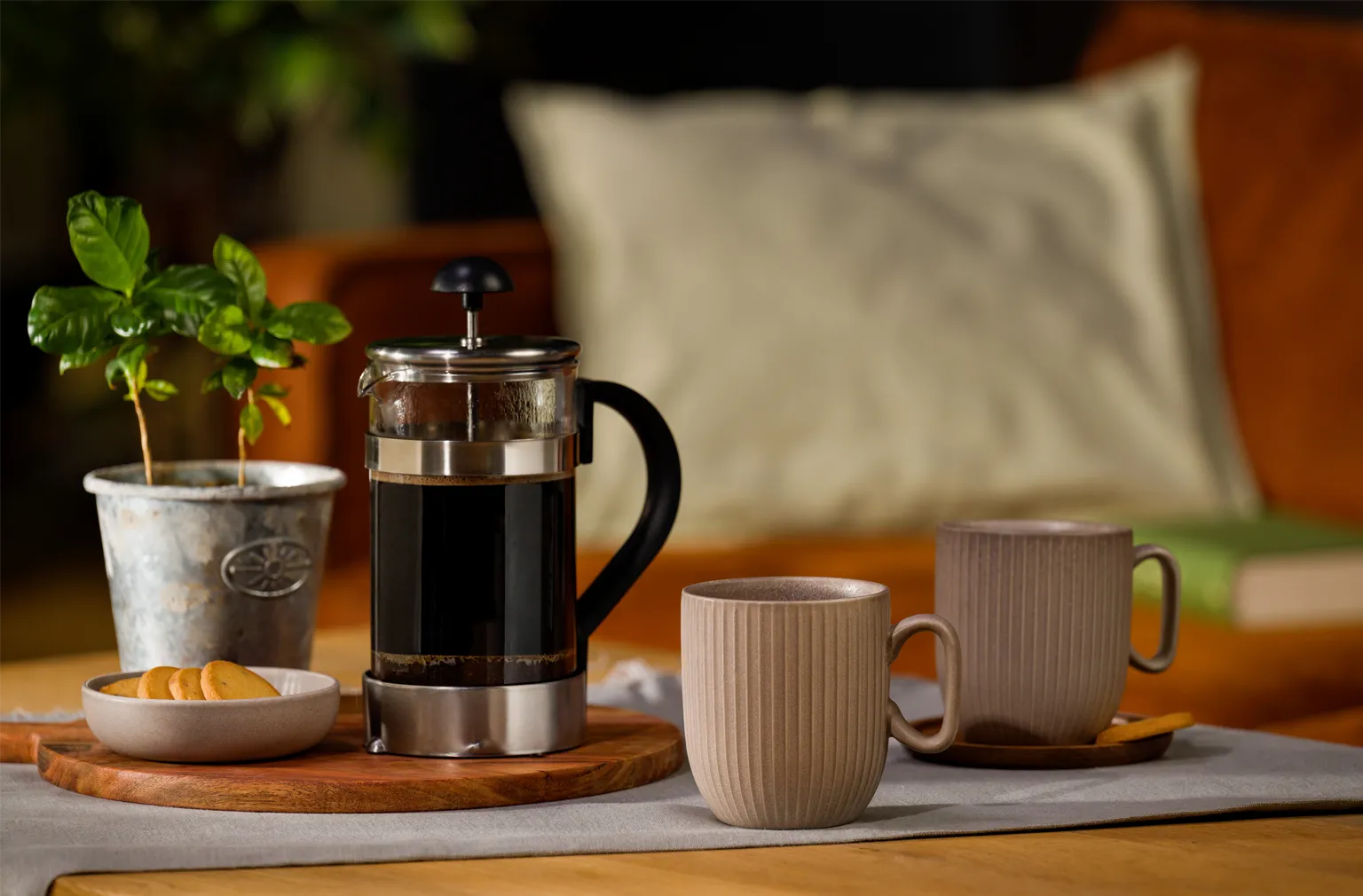 Zwei beige Tassen stehen neben einer gefüllten French Press auf einem gedeckten Tisch, daneben ein kleiner Keks­teller und eine Topfpflanze
