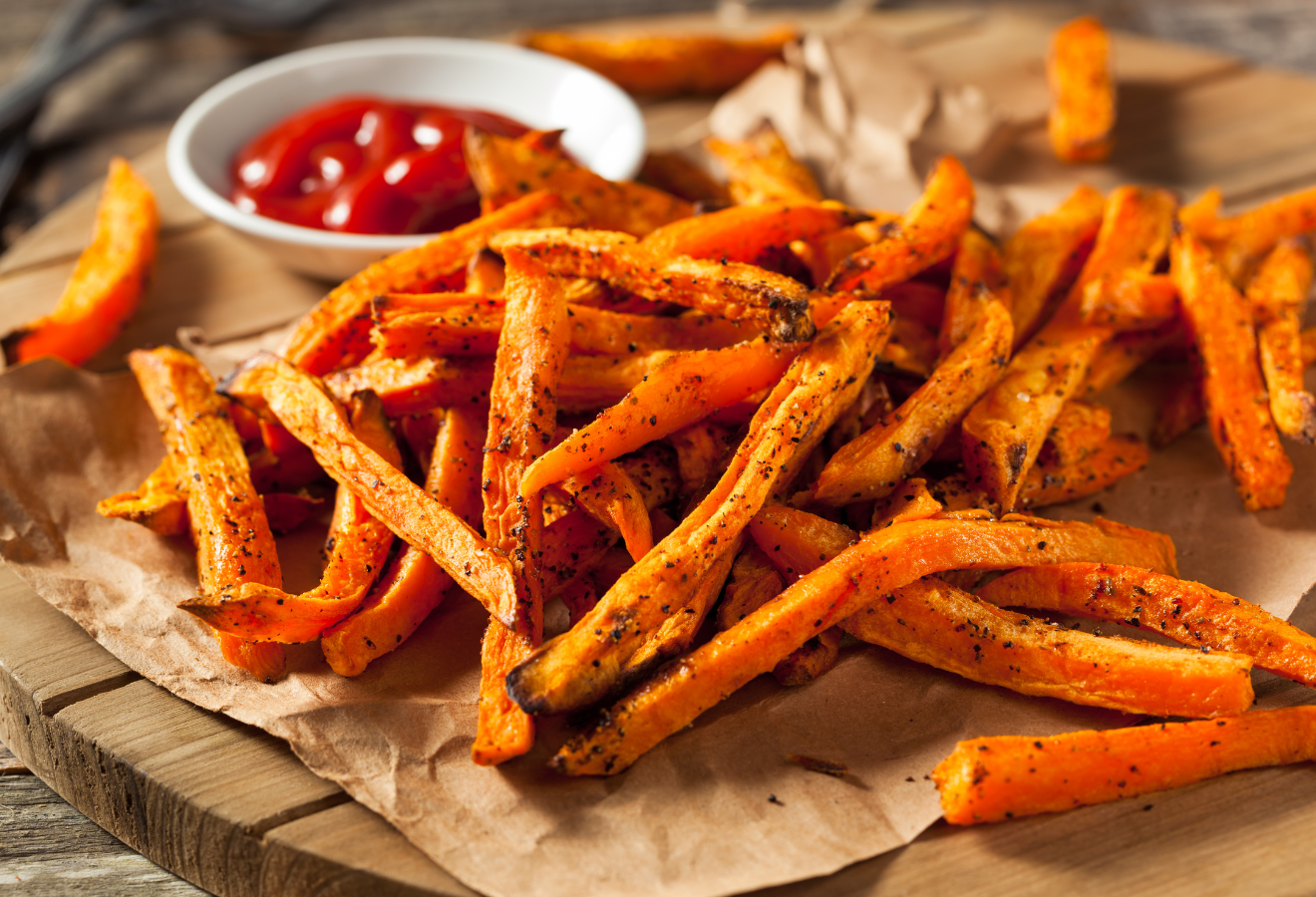Süßkartoffelpommes vom Blech auf einem Holzbrett mit Backpapier im Hintergrund eine weiße Schale mit Ketchup darin