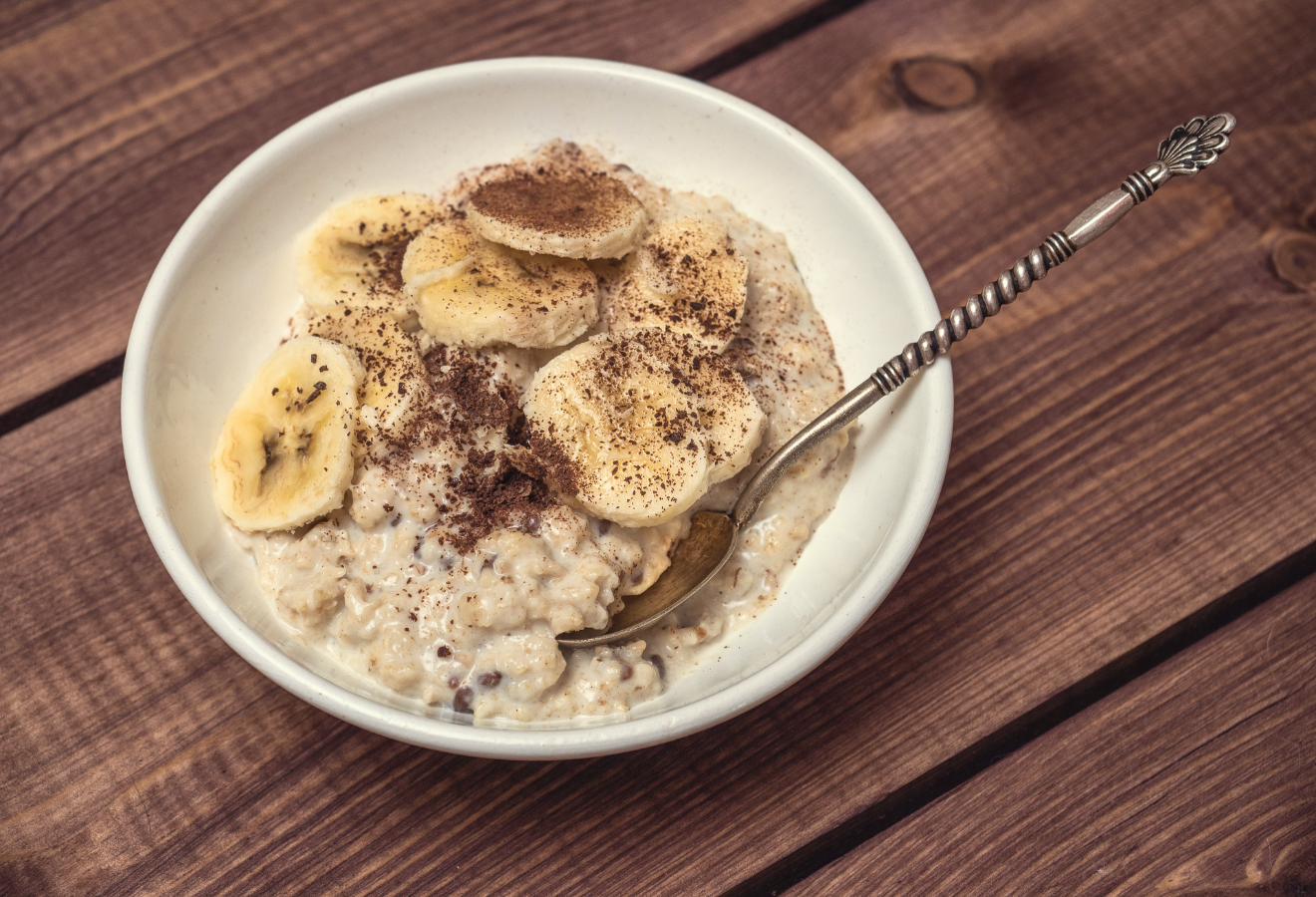 Veganes Knusper-Schoko-Porridge mit Banane in einer weißen Schüssel mit Kakaopulver garniert und einem Löffel darin