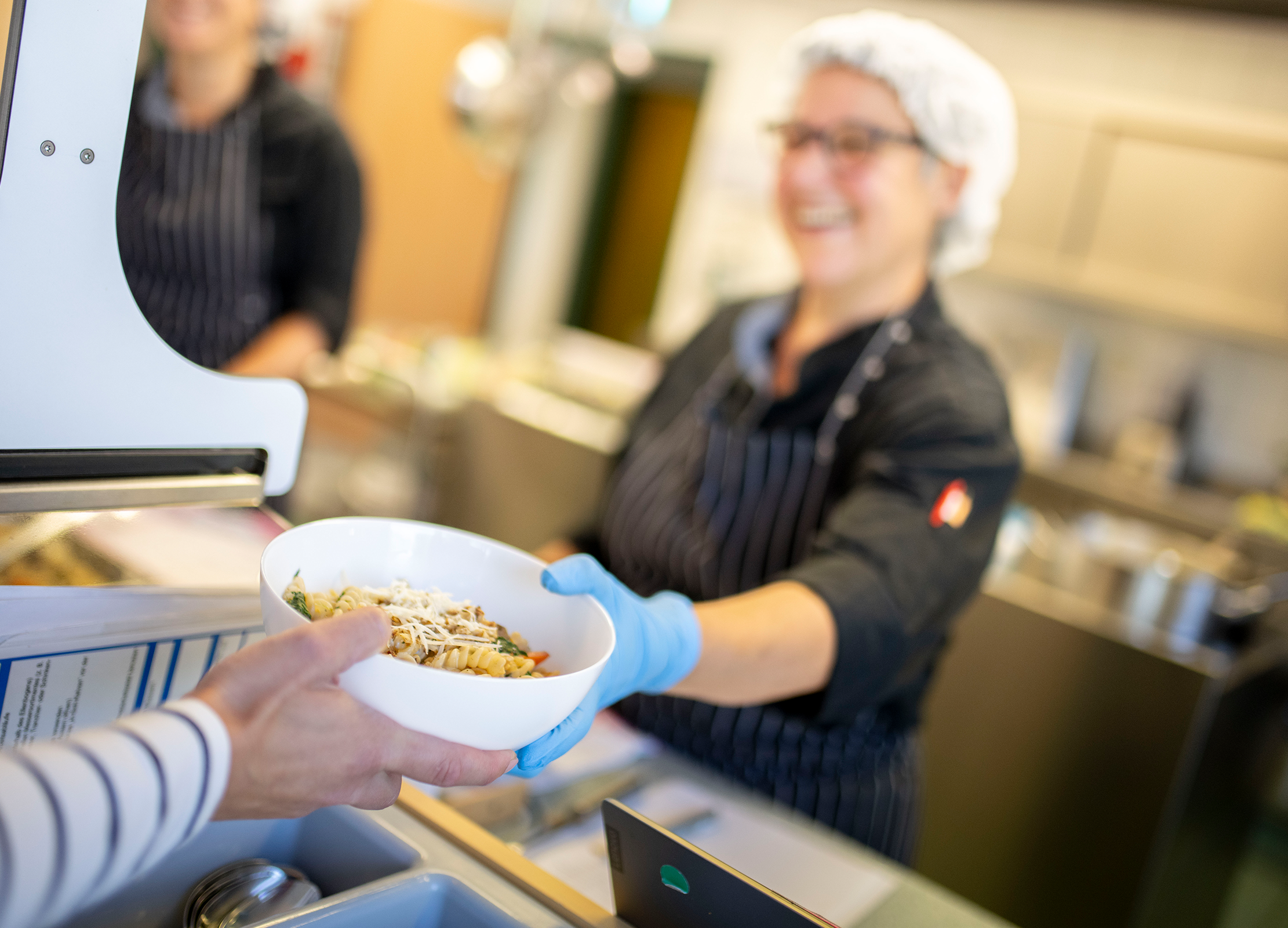 Küchenmitarbeiterin reicht eine Schale mit Pasta über die Essensausgabe