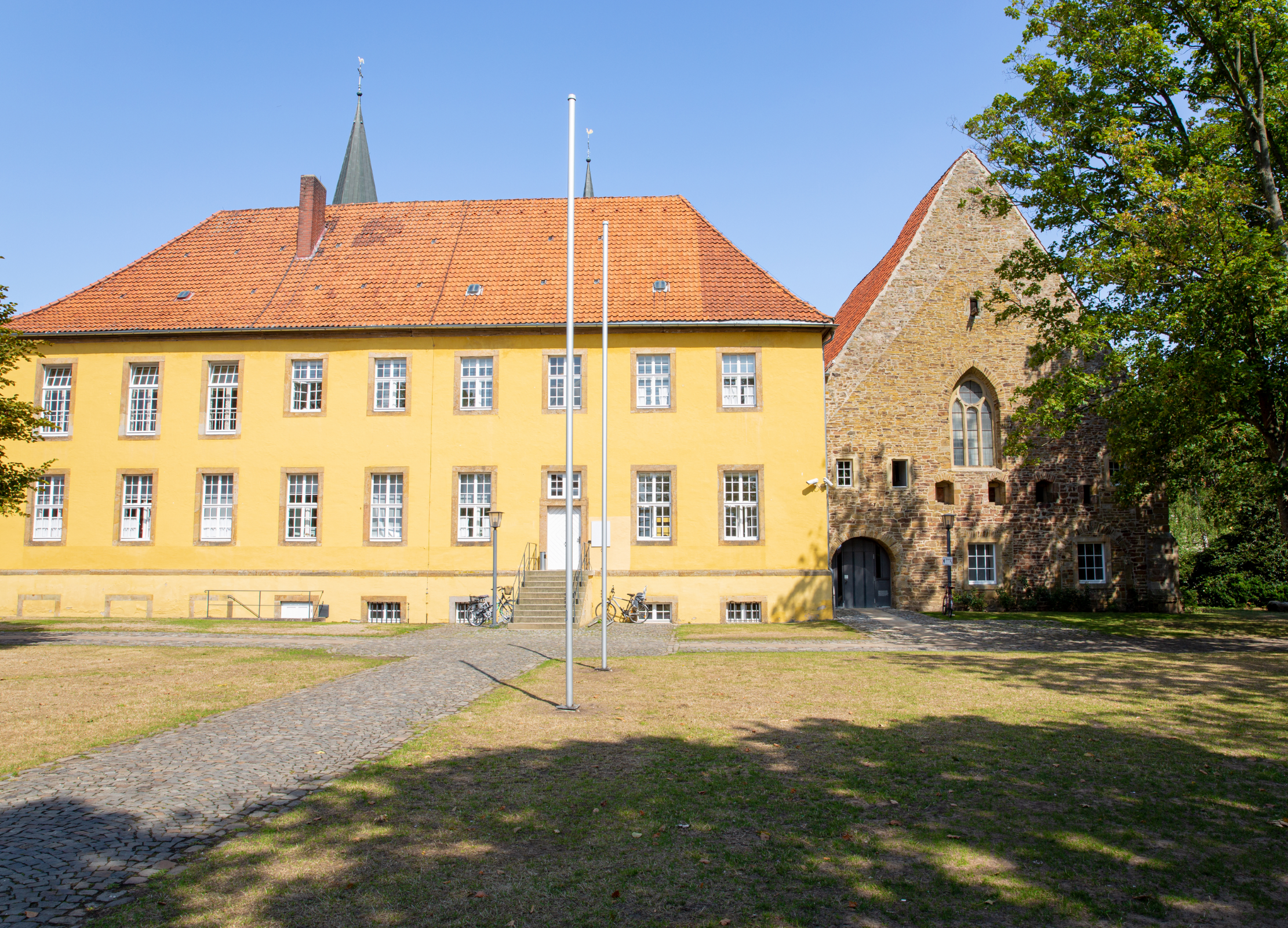 Gelbes historisches Gebäude mit rotem Dach und angrenzender Backsteinkirche auf einem Platz mit Fahnenmasten