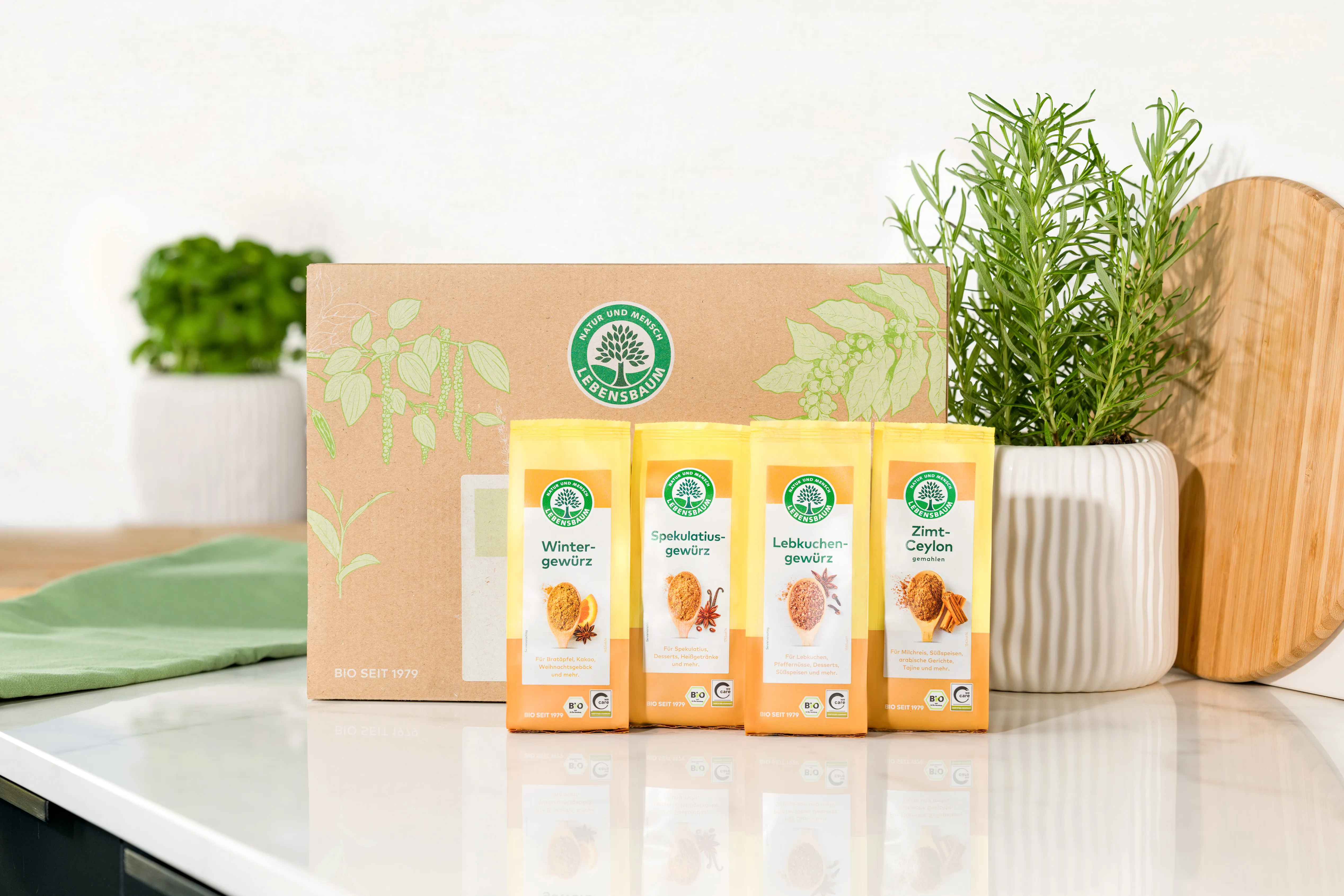 Lebensbaum Bio-Gewürzset mit vier Sorten in orangefarbenen Päckchen: Wintergewürz, Spekulatiusgewürz, Lebkuchengewürz und Zimt-Ceylon vor Kraft-Karton, umgeben von Kräutertöpfen und Holzaccessoires.