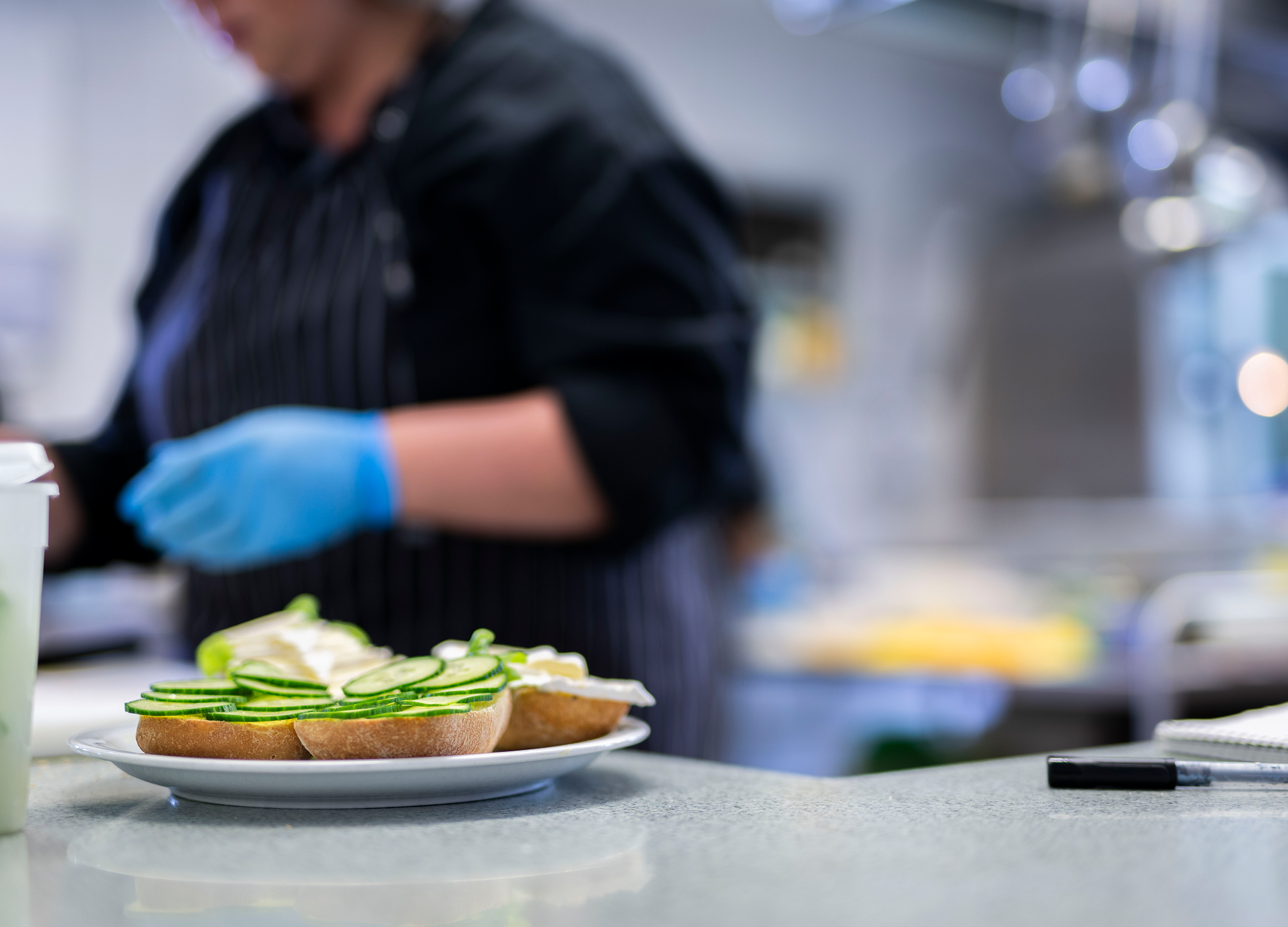 Küchenmitarbeiterin bereitet belegte Brötchen mit Gurkenscheiben vor, im Vordergrund ein Teller auf der Theke