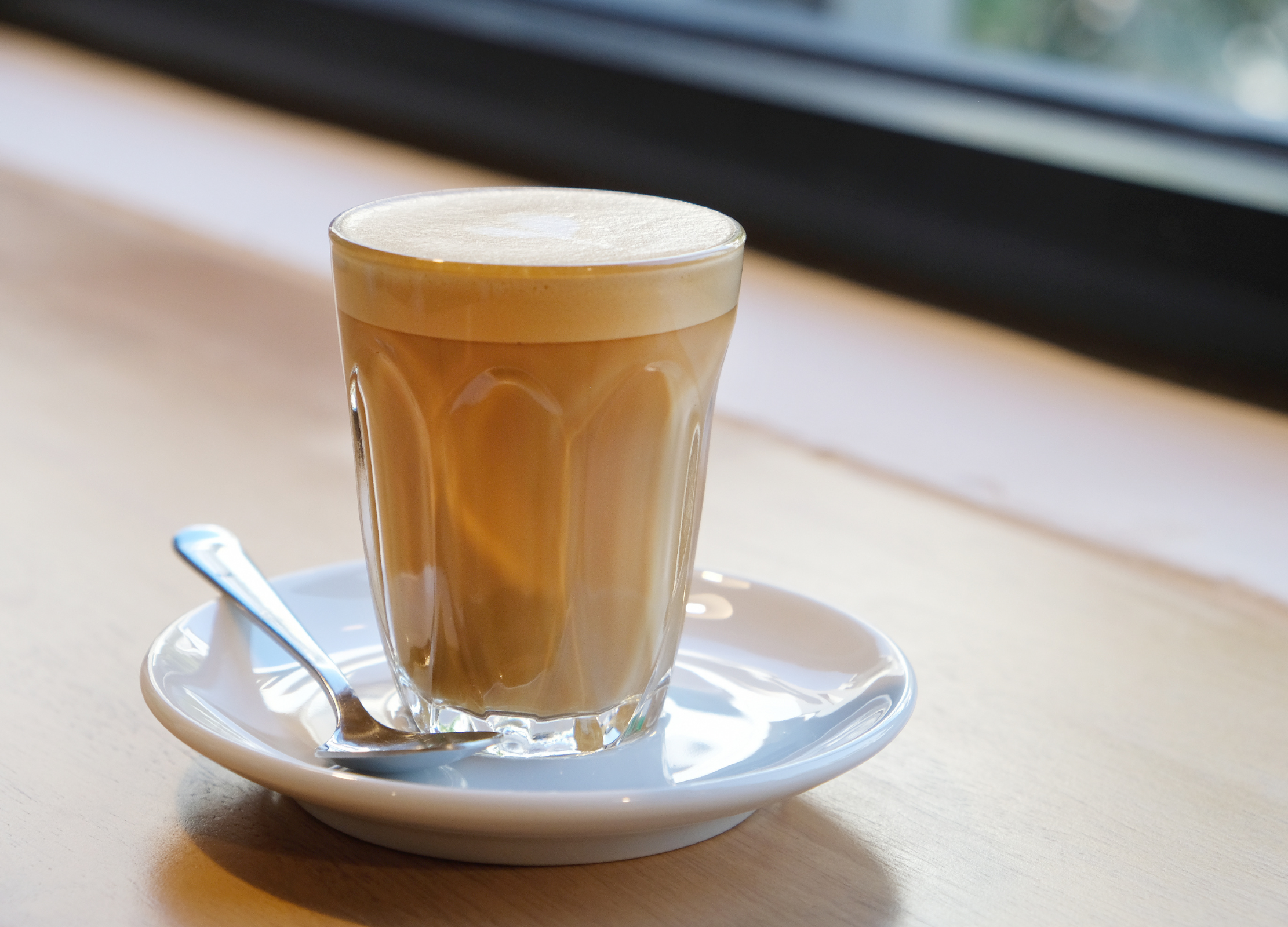 Ein Glas mit cremigem Milchkaffee steht auf einem weißen Unterteller mit Löffel auf einer Holzoberfläche