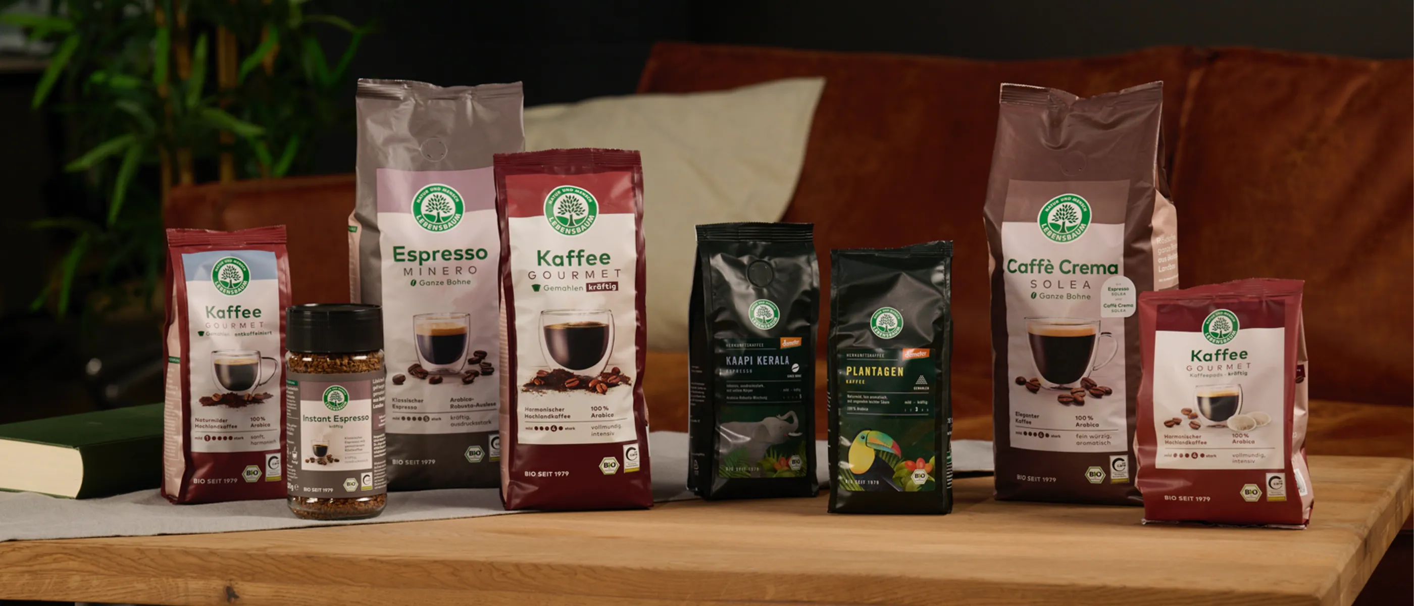 Verschiedene Kaffeeprodukte von Lebensbaum sind auf einem Holztisch arrangiert, im Hintergrund ein braunes Sofa und grüne Pflanzen