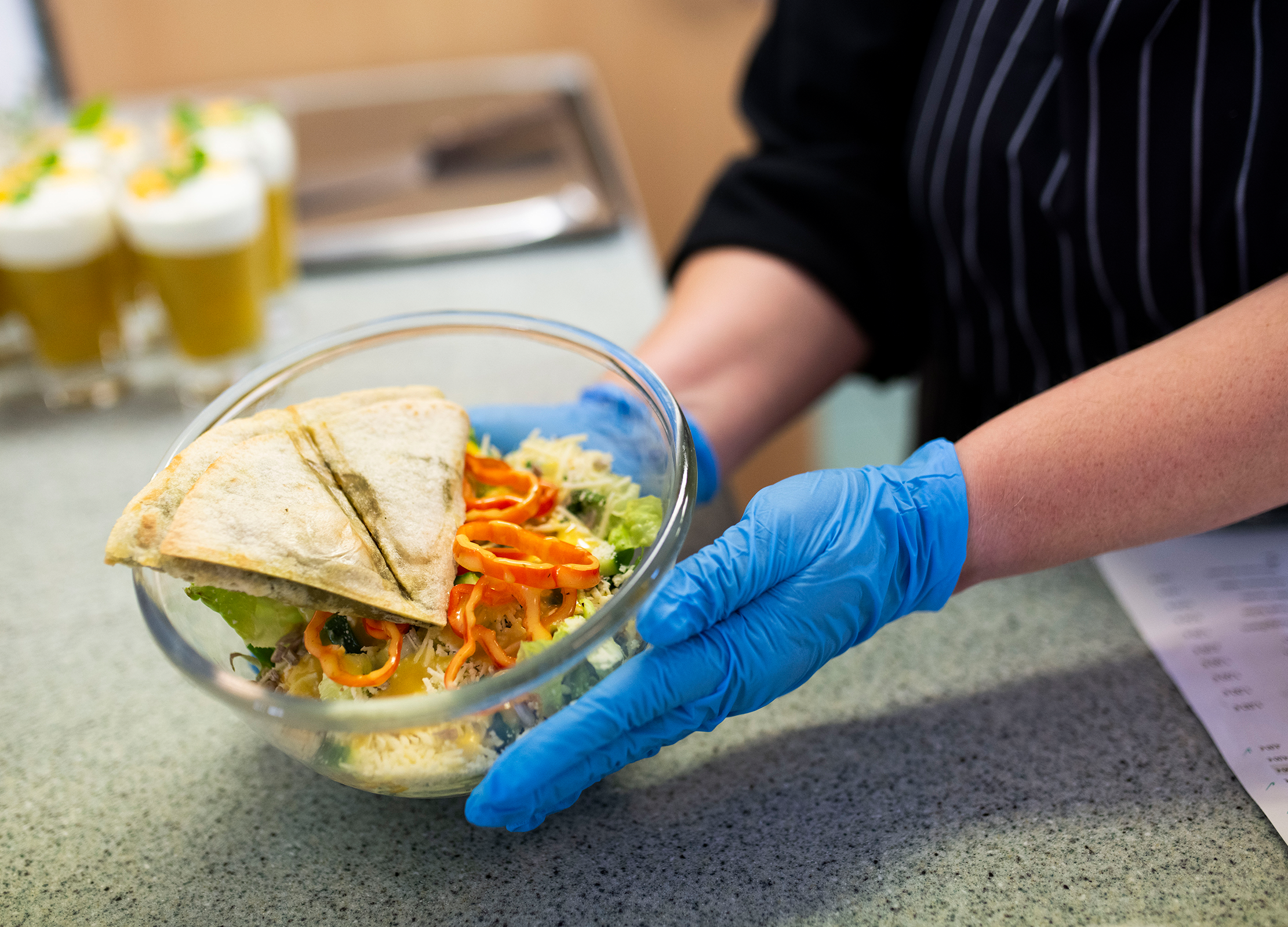 Person mit blauen Handschuhen hält eine Glasschale mit Salat, Gemüse und einem gefalteten Fladenbrot