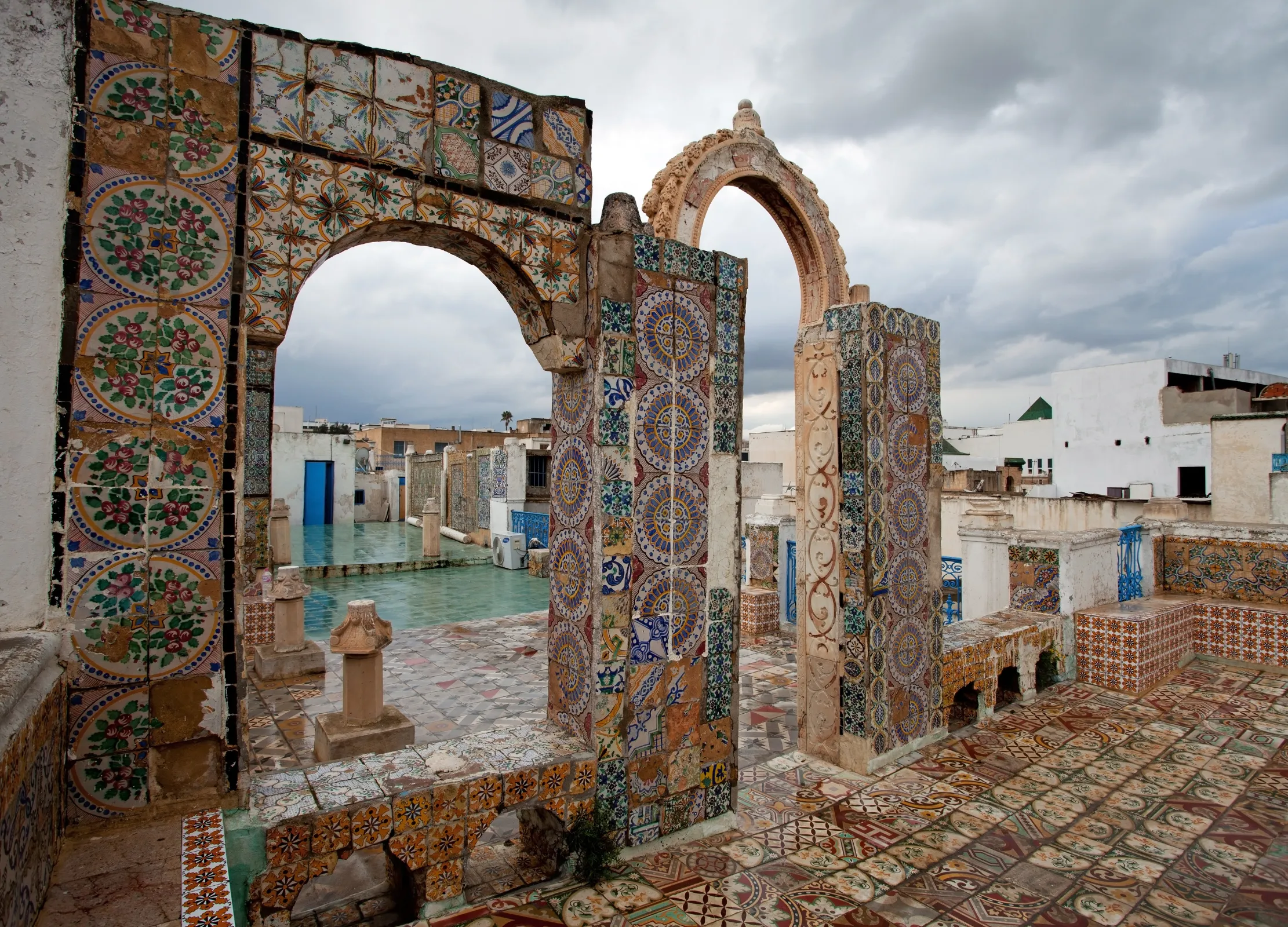 Bunt verzierte Torbögen und Mosaikfliesen schmücken einen historischen Innenhof unter bewölktem Himmel in Tunesien