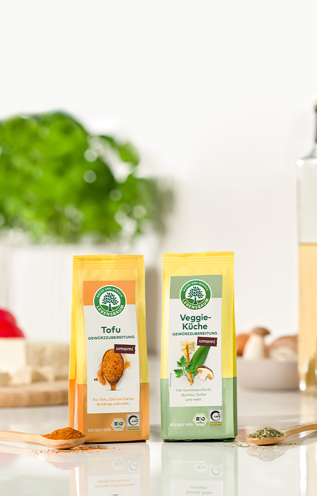 Zwei Gewürzpackungen mit der Aufschrift ‚Tofu‘ und ‚Veggie-Küche‘, daneben passende Gewürze auf Holzlöffeln