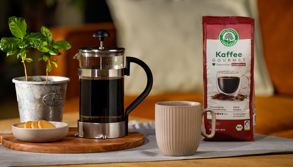 Packung Lebensbaum Kaffee Gourmet steht neben einer French Press und einer Tasse auf einem gedeckten Tisch, daneben ein kleiner Keks­teller und eine Kaffeepflanze im Topf