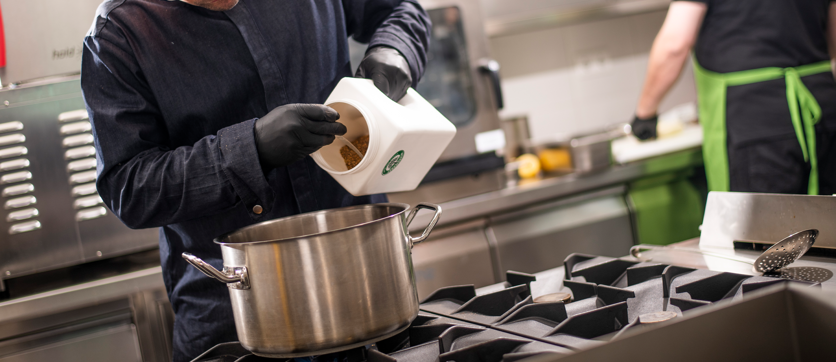 Person schüttet Gewürze aus einer Lebensbaum-Dose in einen großen Kochtopf in einer Profiküche