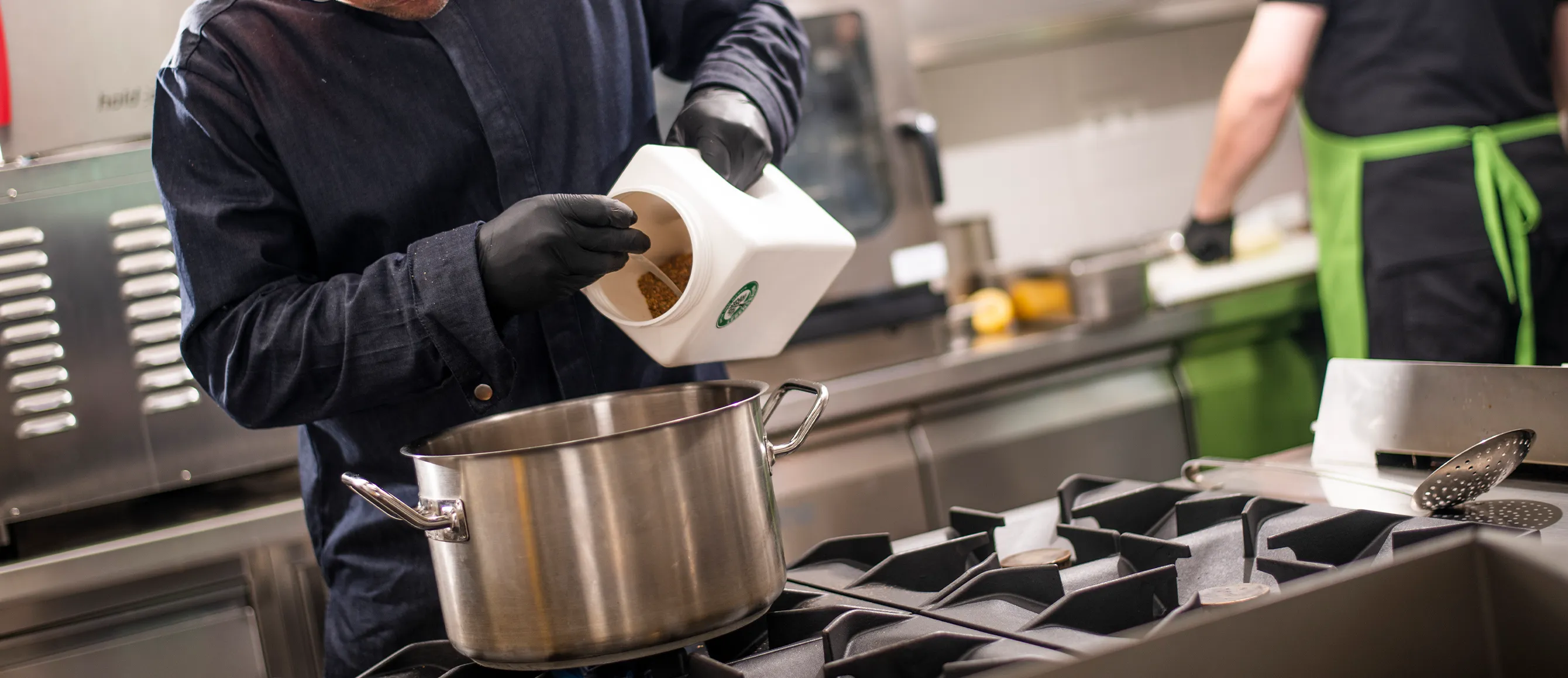 Person schüttet Gewürze aus einer Lebensbaum-Dose in einen großen Kochtopf in einer Profiküche