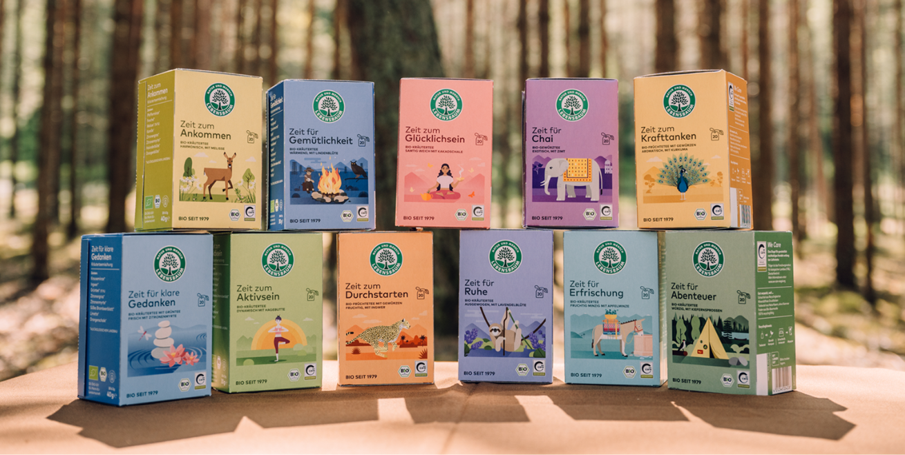 Auf einem Holztisch sind verschiedene Teepackungen aufgereiht und teils übereinander gestapelt. Im Hintergrund ist ein Wald zu sehen