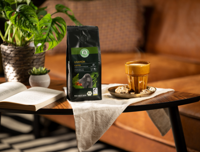 Kaffeepackung „Uganda“ mit Gorilla-Motiv steht neben einer dampfenden Tasse und Gebäck auf einem Tisch vor einem Ledersofa