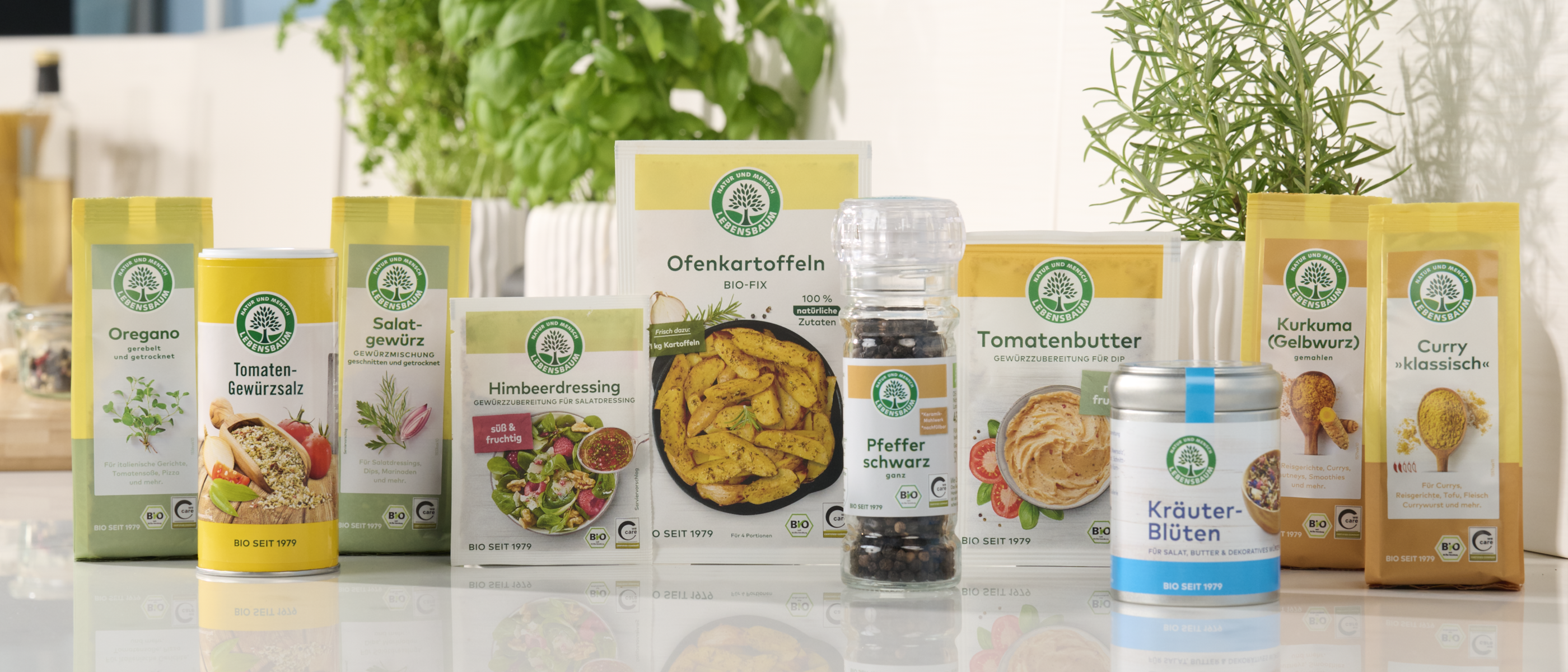 Mehrere Gewürz- und Kräuterprodukte von Lebensbaum stehen nebeneinander auf einer Küchenarbeitsfläche, im Hintergrund sind frische Kräuterpflanzen zu sehen