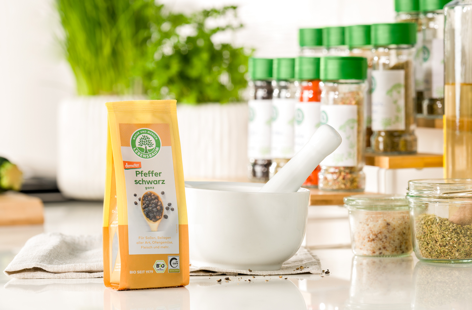 Packung Lebensbaum Pfeffer schwarz steht neben einem weißen Mörser auf einer hellen Küchenarbeitsfläche, im Hintergrund frische Kräuter und Gewürze
