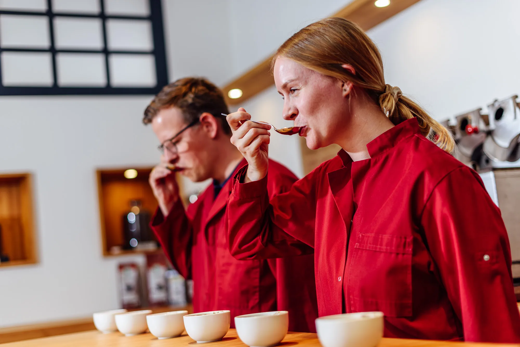 Zwei Verkoster in roten Uniformen probieren Kaffee-Sorten aus weißen Tassen während einer professionellen Verkostung in moderner Rösterei.