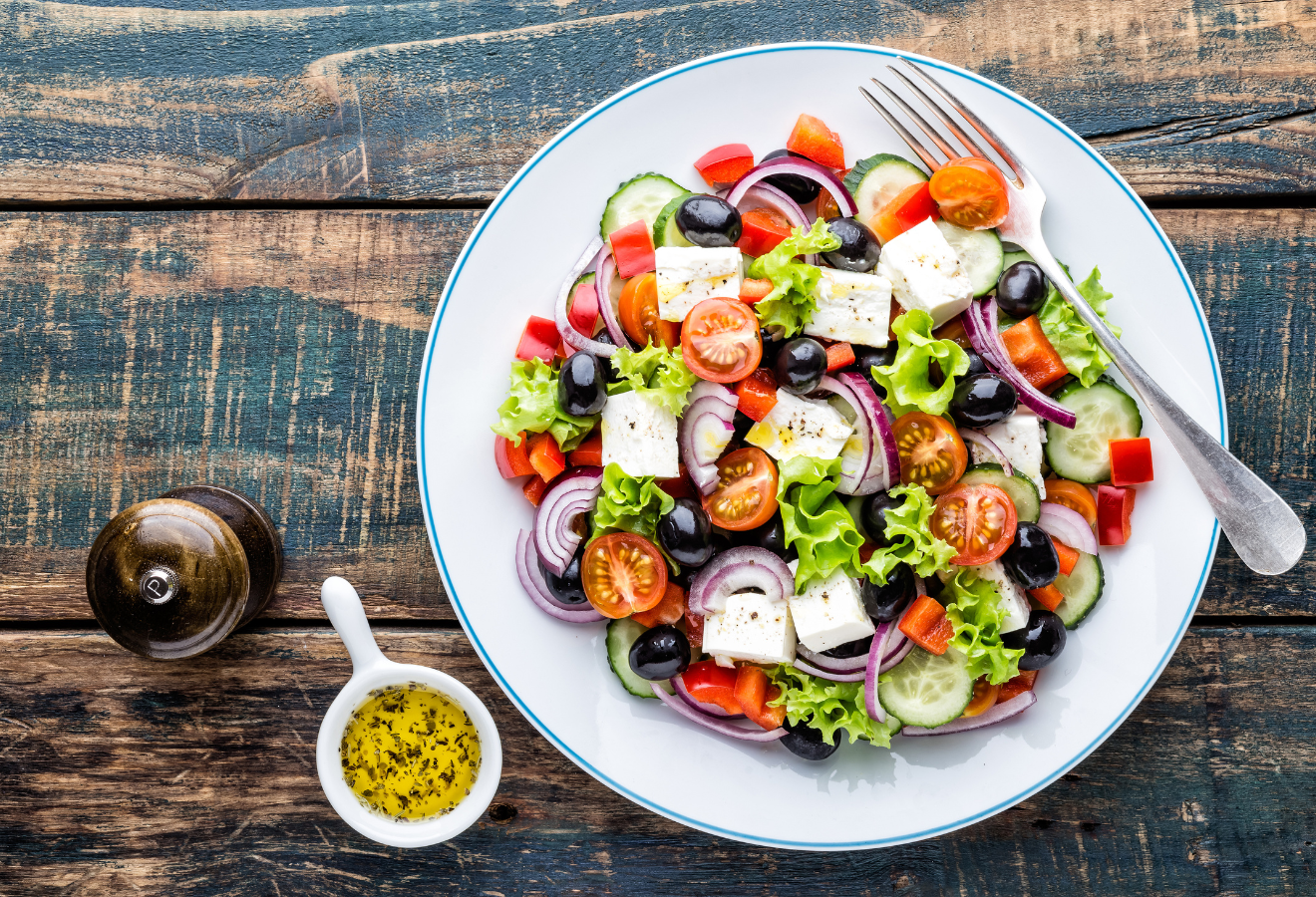 Griechischer Salat auf einem weißen Teller mit einer Gabel, daneben Dressing in einem Schälchen
