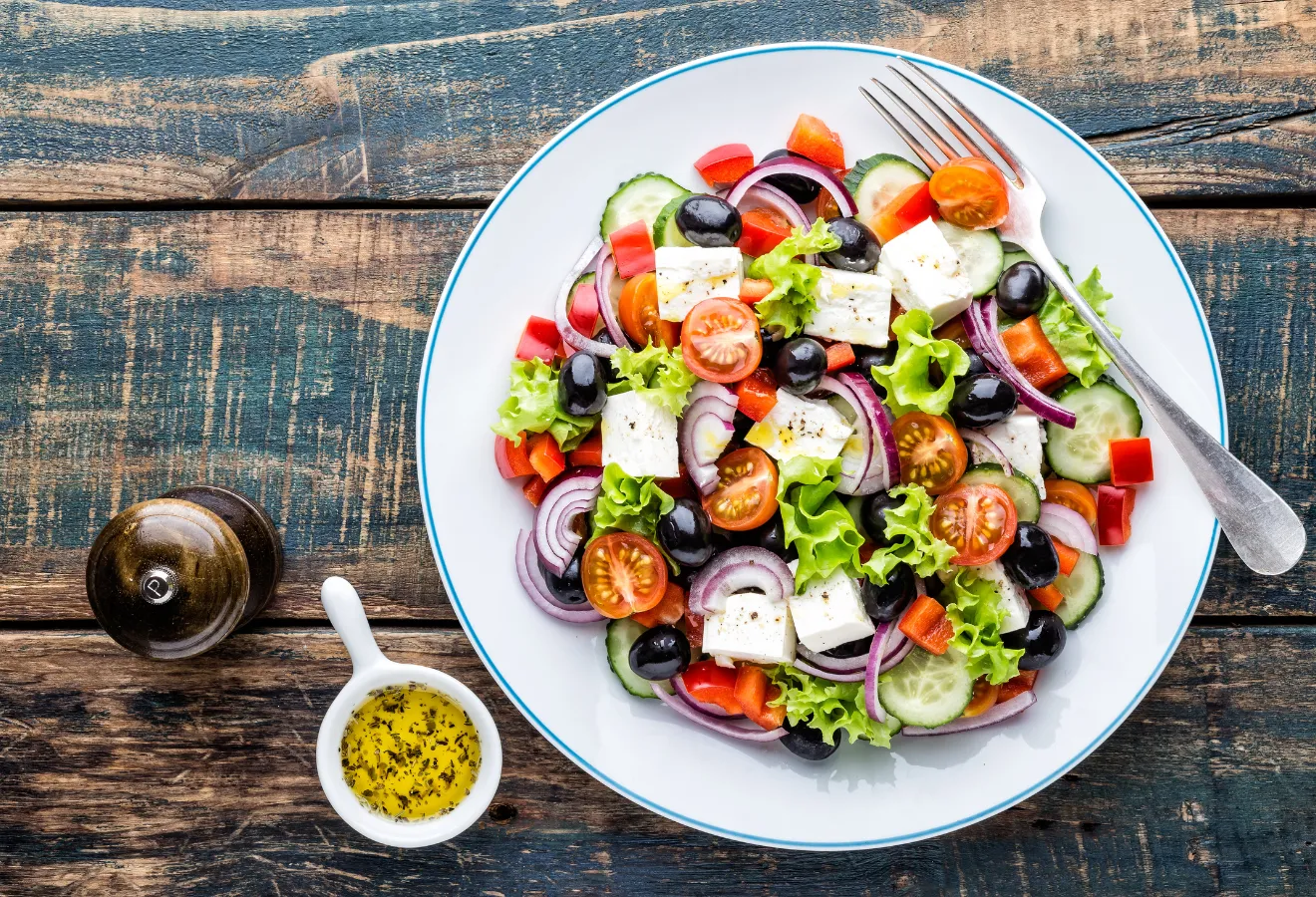 Griechischer Salat auf einem weißen Teller mit einer Gabel, daneben Dressing in einem Schälchen