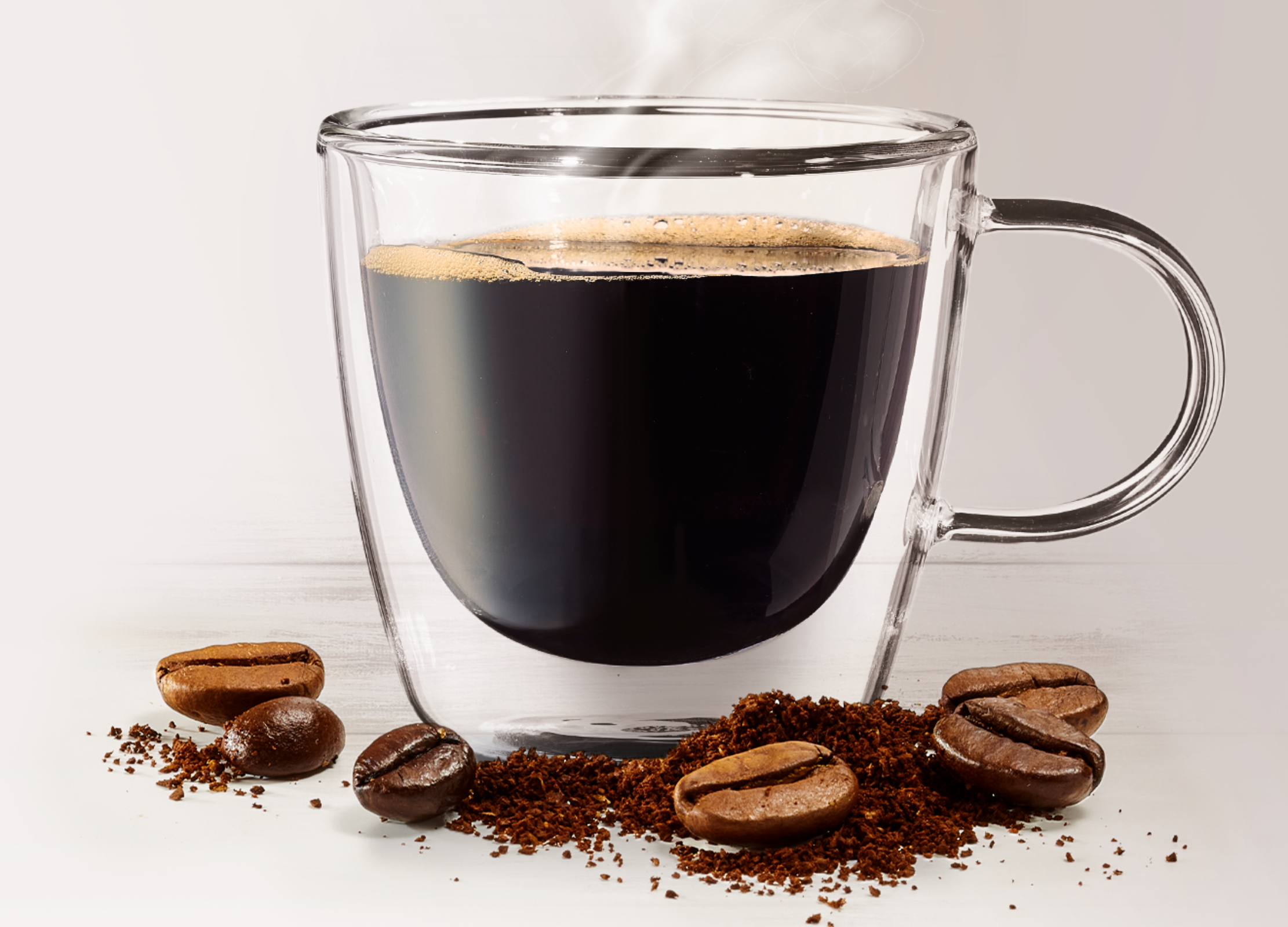 Eine Tasse schwarzer Kaffee dampft in einem Glasbecher, umgeben von Kaffeebohnen und Kaffeepulver