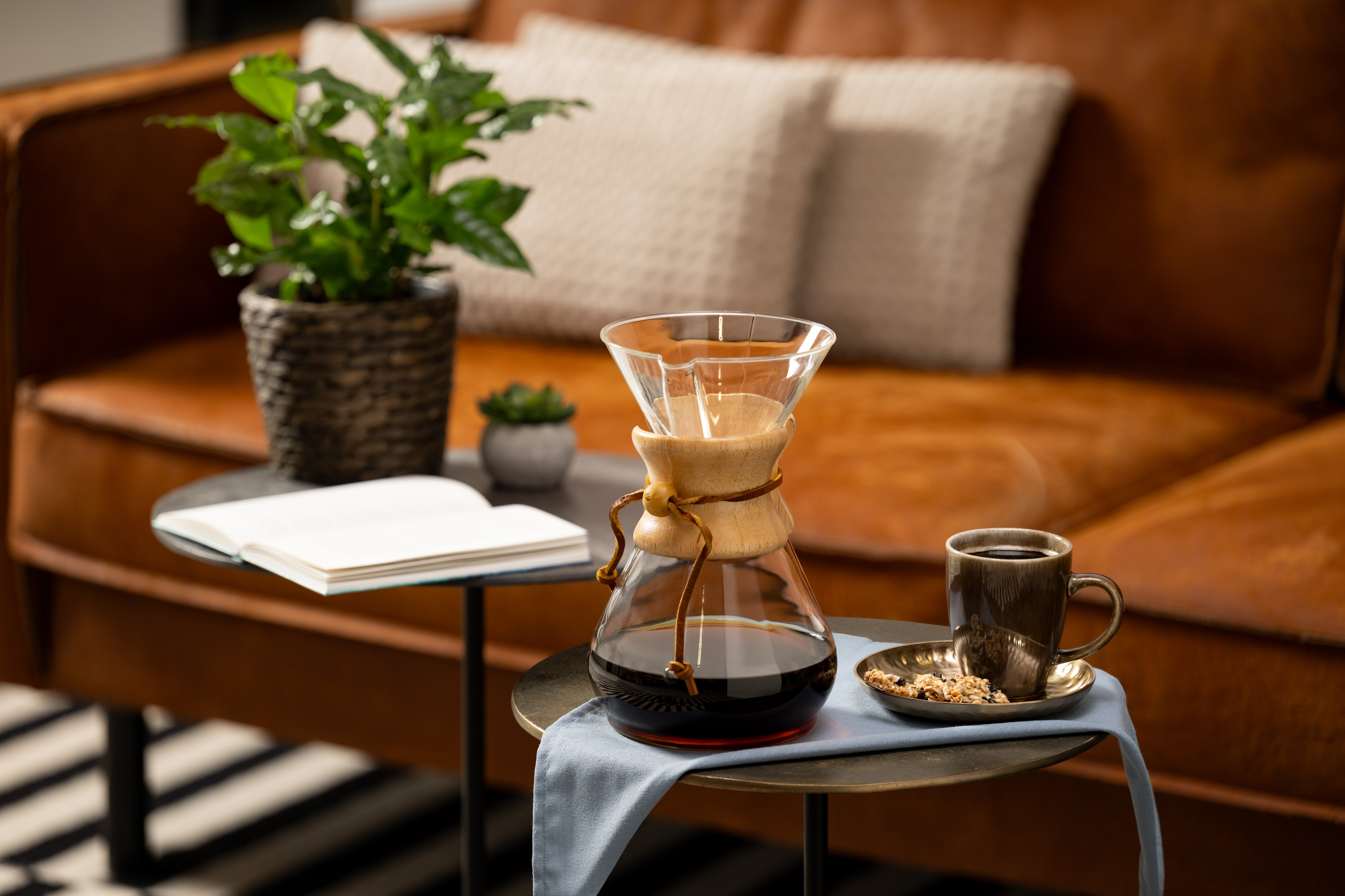 Chemex-Kaffeezubereiter mit frisch gebrühtem dunklem Kaffee, daneben Tasse mit Kaffee und Gebäck in gemütlichem Wohnzimmer mit Sofa.