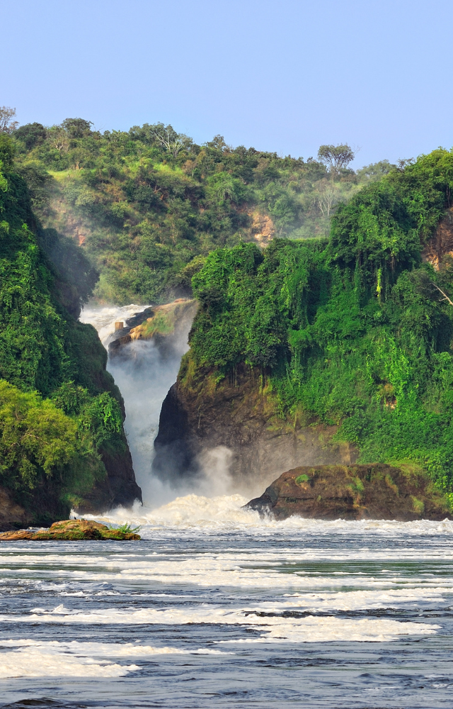 Wasserfall stürzt zwischen grünen Felsen in einen breiten Fluss in Uganda