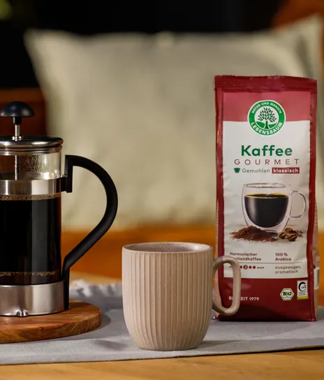Packung Lebensbaum Kaffee Gourmet steht neben einer French Press und einer Tasse auf einem gedeckten Tisch, daneben ein kleiner Keks­teller und eine Kaffeepflanze im Topf