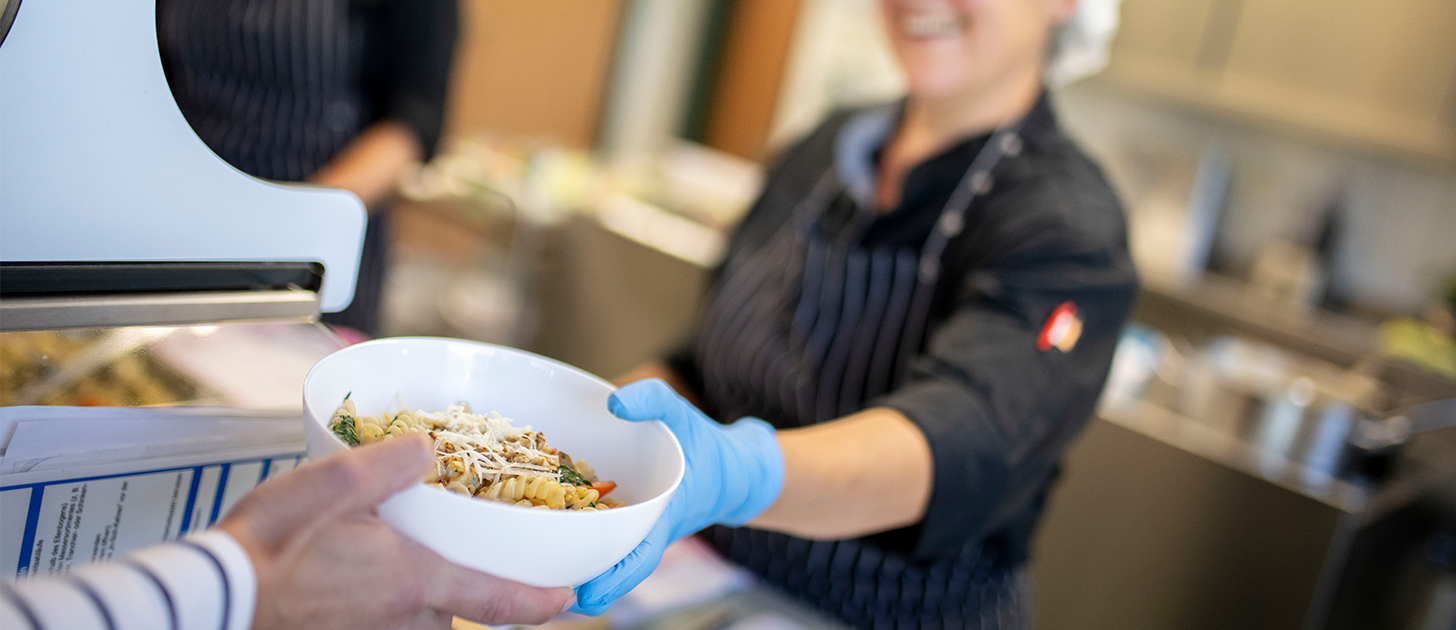 Küchenmitarbeiterin reicht eine Schale mit Pasta über die Essensausgabe
