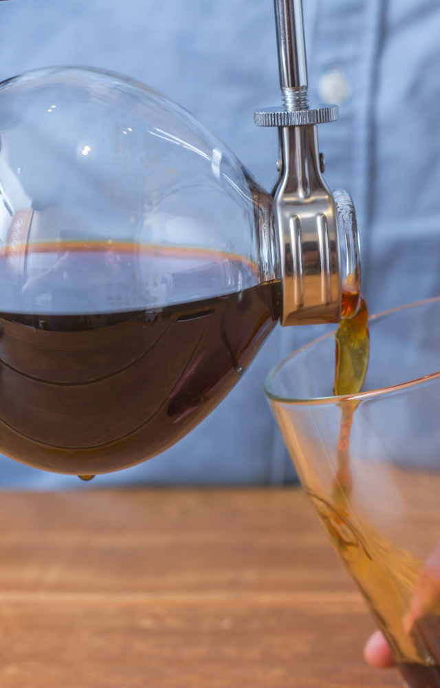 Frisch gebrühter Kaffee wird aus der Glaskammer eines Syphon-Kaffeebereiters in ein Glas eingeschenkt