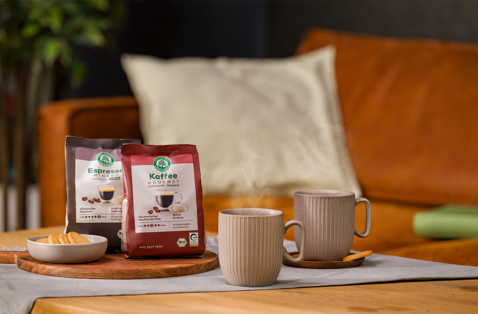 Zwei dampfende Tassen Kaffee stehen auf einem gedeckten Tisch neben einer Keks­schale und zwei Packungen Lebensbaum Kaffee und Espresso