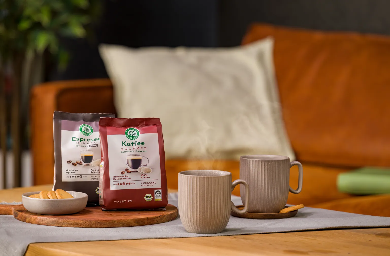 Zwei dampfende Tassen Kaffee stehen auf einem gedeckten Tisch neben einer Keks­schale und zwei Packungen Lebensbaum Kaffee und Espresso