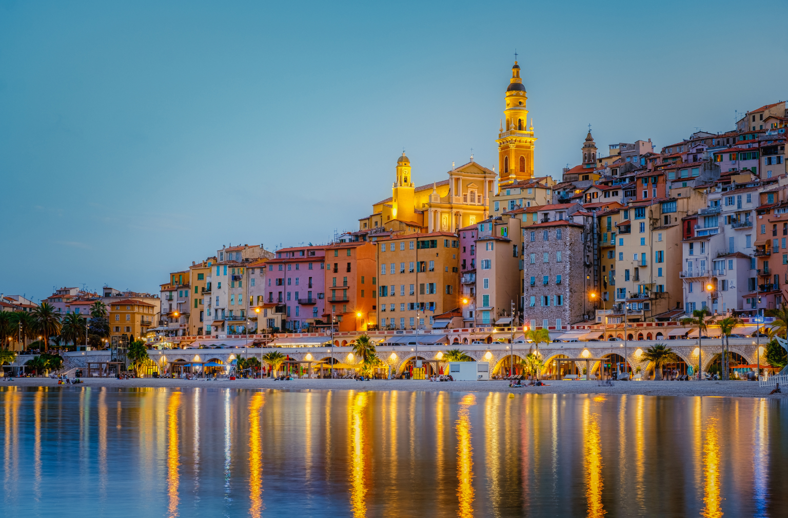 Bunte Häuser und beleuchtete Kirche an einer Promenade spiegeln sich im Wasser bei Abenddämmerung