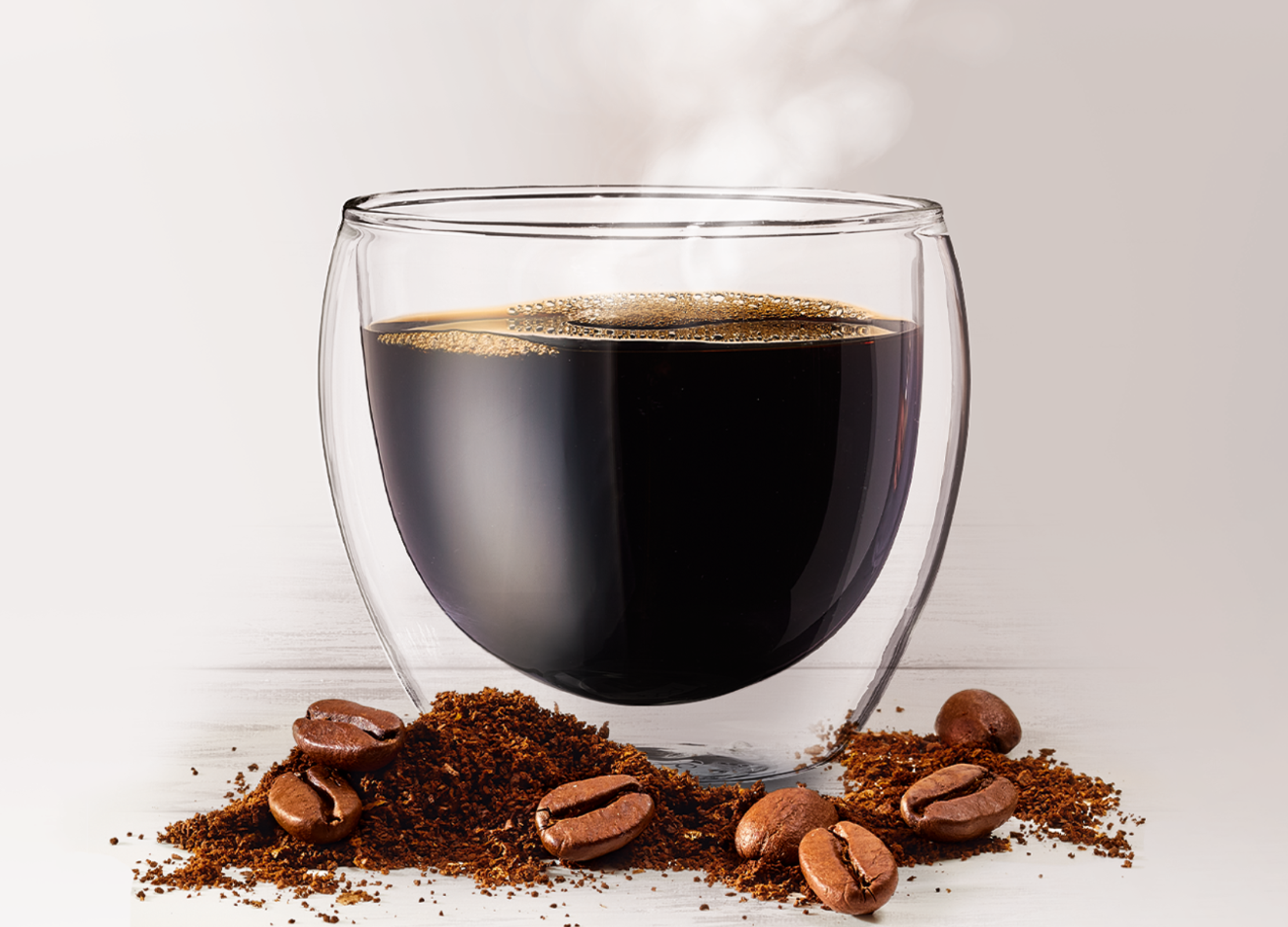 Eine Tasse schwarzer Kaffee steht dampfend inmitten von Kaffeebohnen und gemahlenem Kaffee auf hellem Untergrund