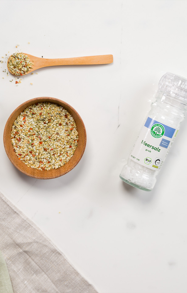 Eine Lebensbaum Salzmischung in der Mühle liegt neben einem Holzlöffel und einer Schale mit Gewürzsalz auf weißer Marmorplatte