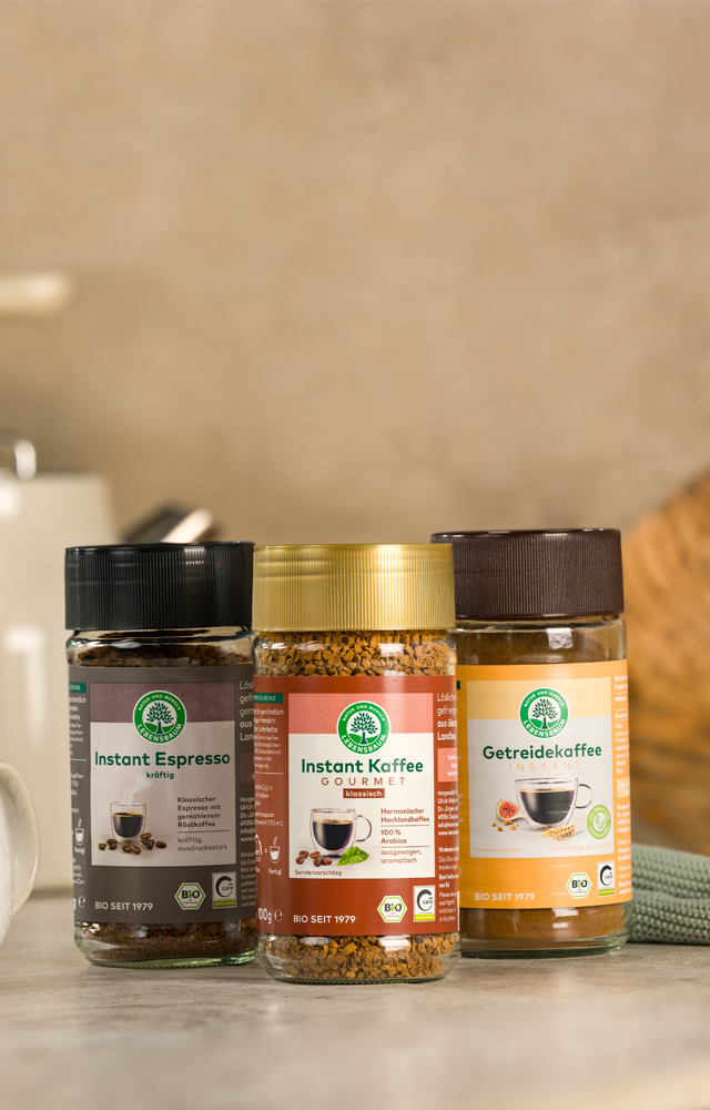 Drei Gläser Instantkaffee von Lebensbaum stehen nebeneinander auf einer Küchenarbeitsfläche, daneben eine Tasse mit Löffel und Küchendeko im Hintergrund