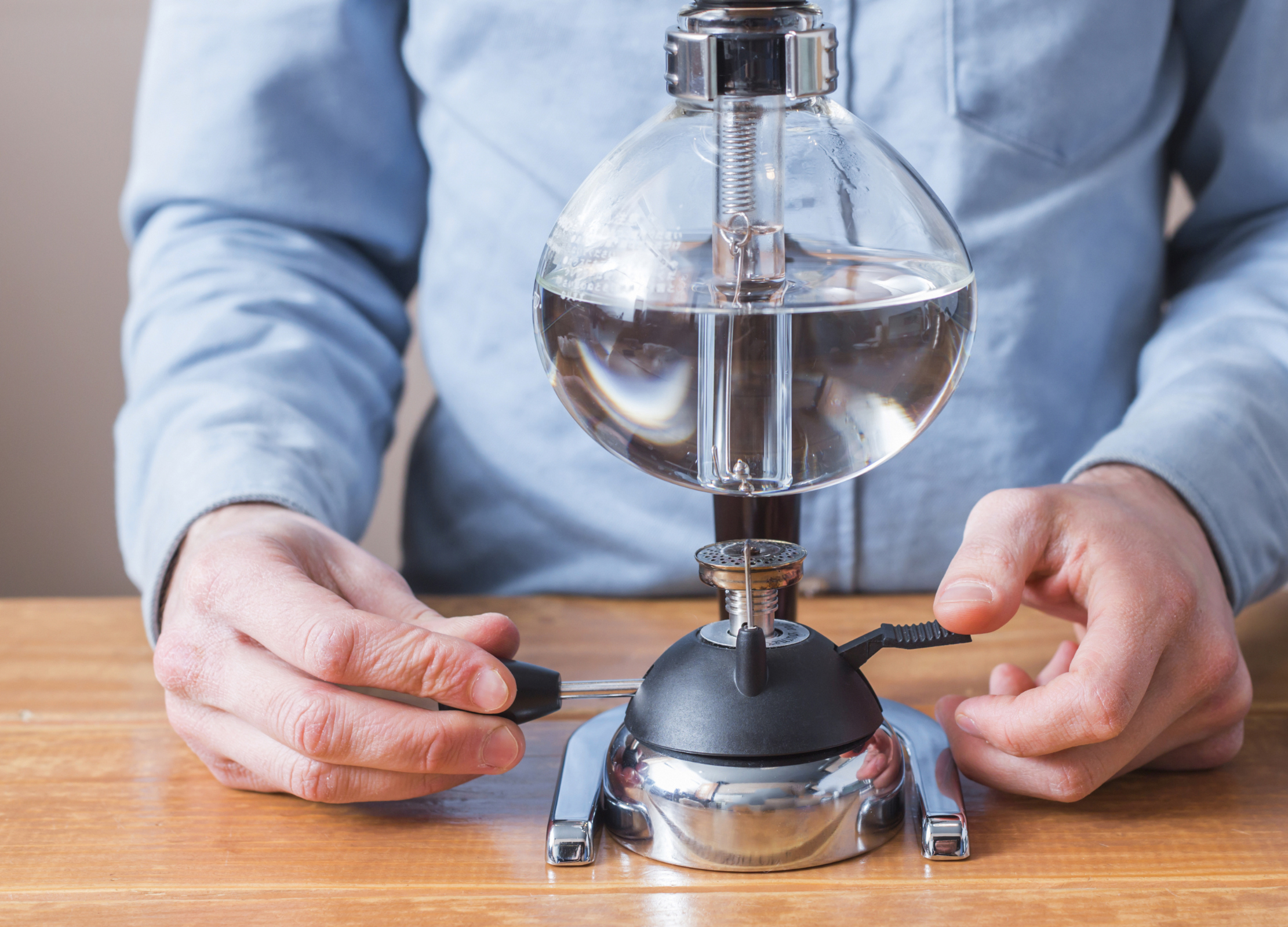 Person bedient einen Syphon-Kaffeebereiter mit rundem Glasbehälter, der auf einem Tisch steht und mit klarem Wasser gefüllt ist
