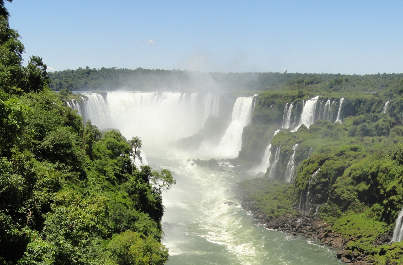 Breite Wasserfälle stürzen zwischen grün bewaldeten Felsen in einen Fluss